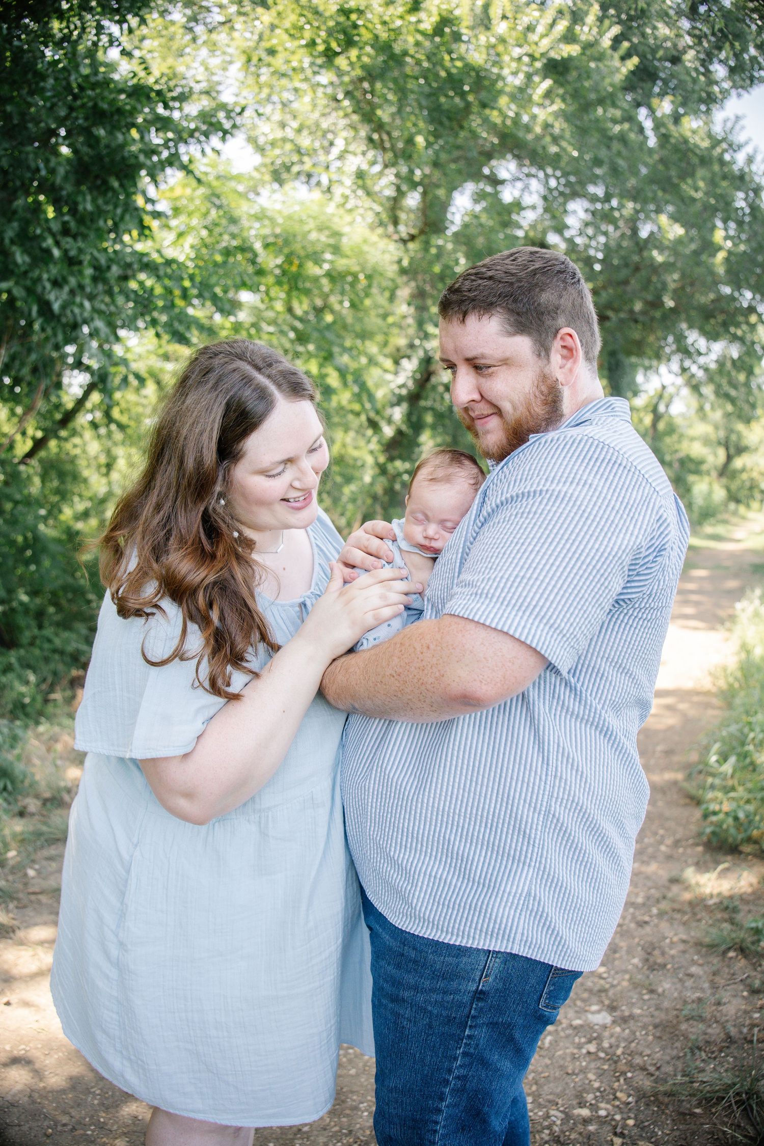 Happy couple smiling at their newborn baby outdoors on a sunny dirt path surrounded by lush green trees.