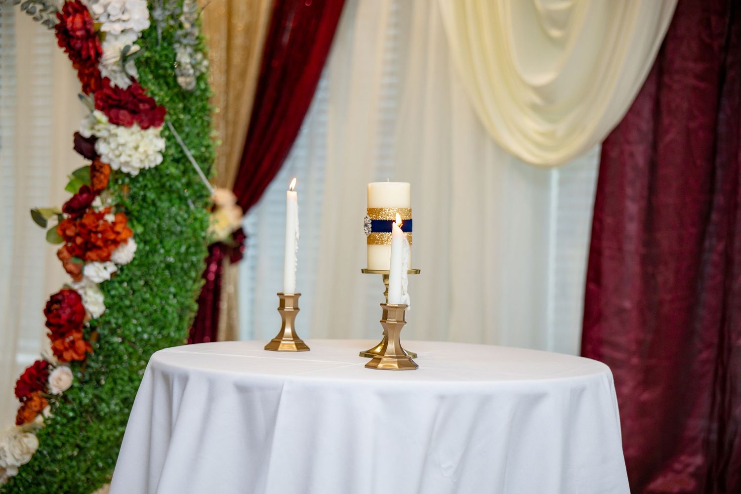Wedding unity candle display on a white draped table with gold candleholders and floral backdrop decorations.