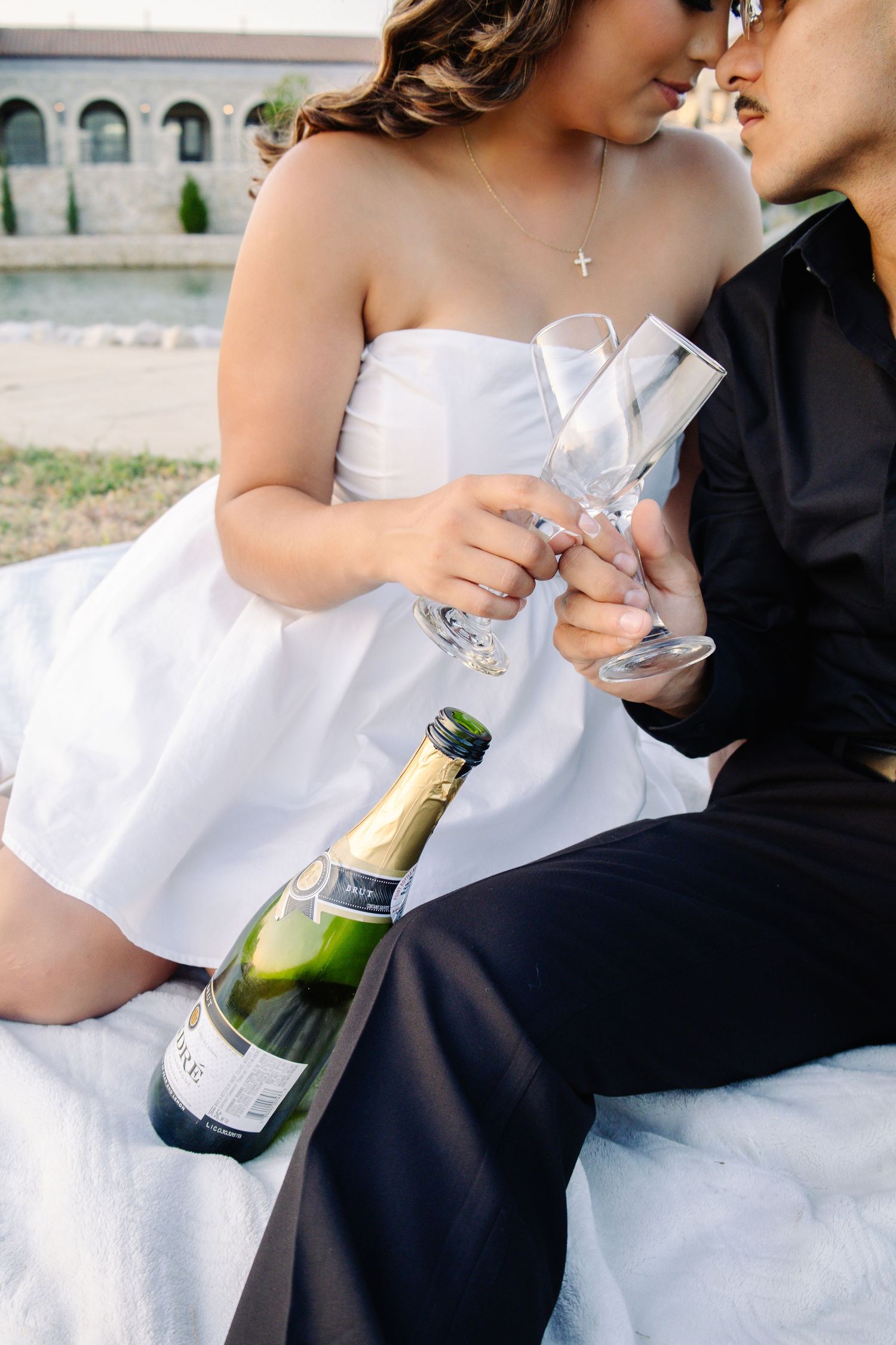 Bride and groom clinking champagne glasses outdoors, with a bottle of sparkling wine resting beside them.