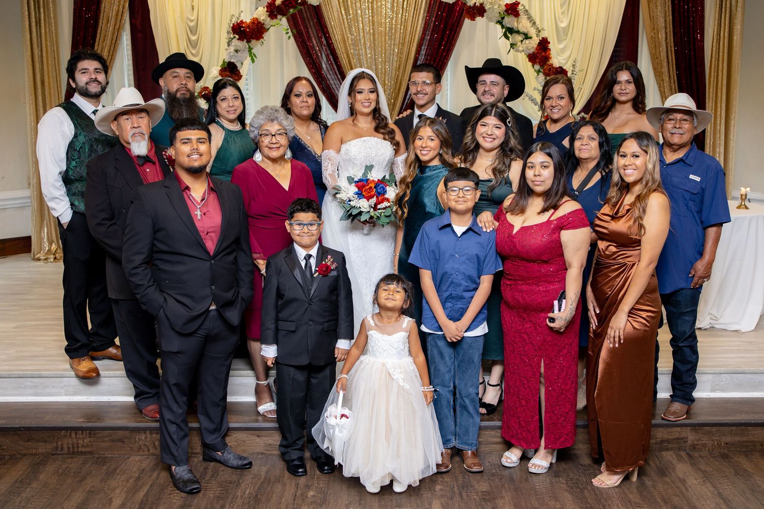 Wedding party group photo with bride, groom, and family in decorated reception hall.
