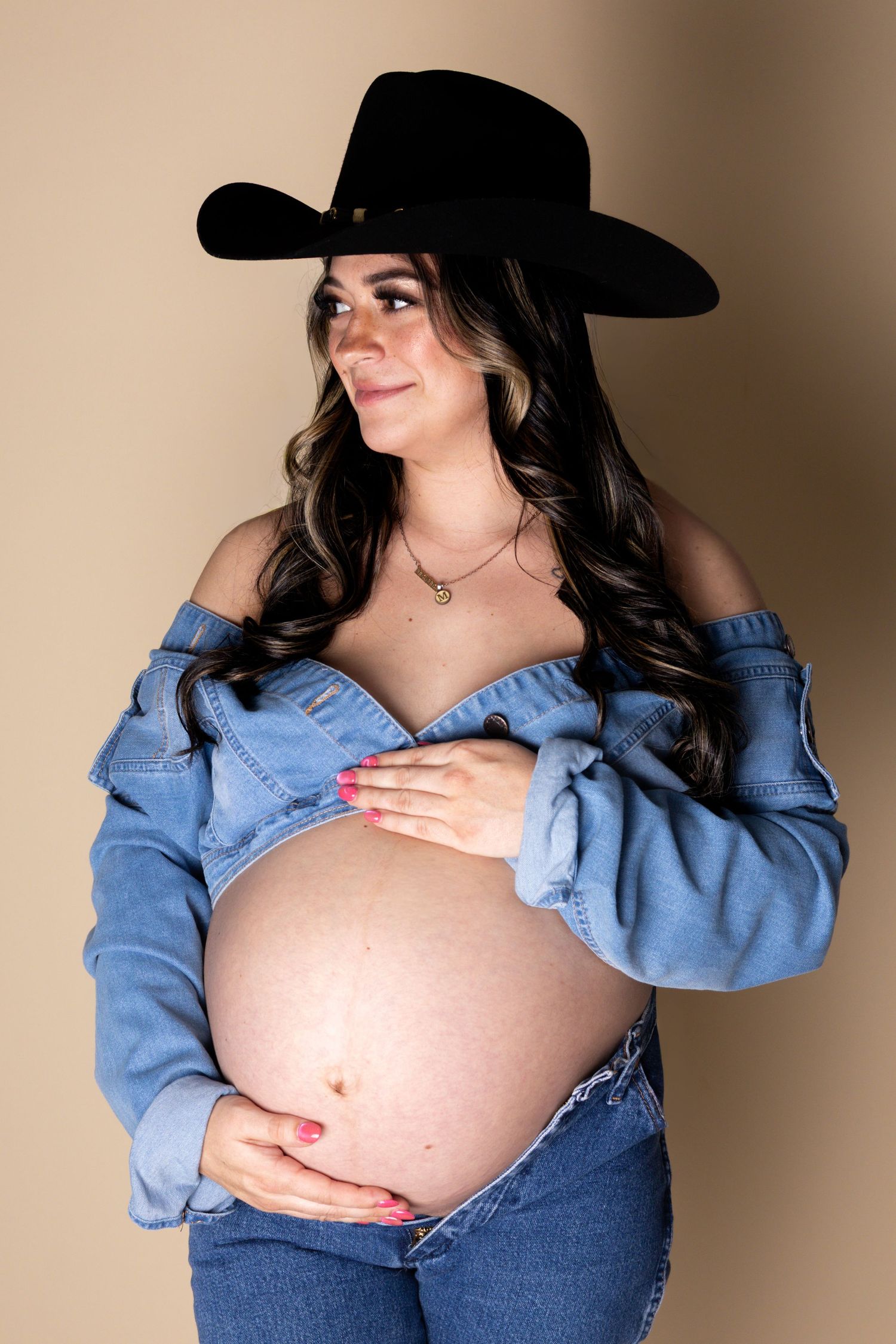 Pregnant woman in black cowboy hat and denim jacket smiling sideways, cradling bare belly.