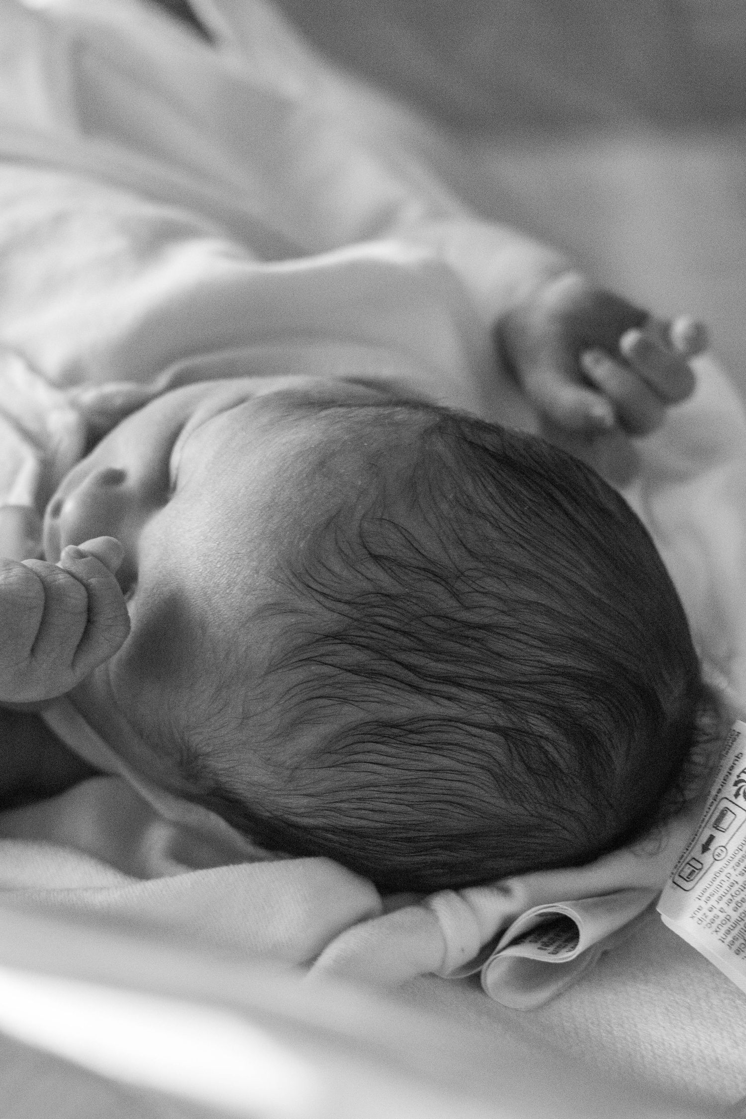 Black and white image of a newborn baby with dark hair lying in a hospital bassinet.