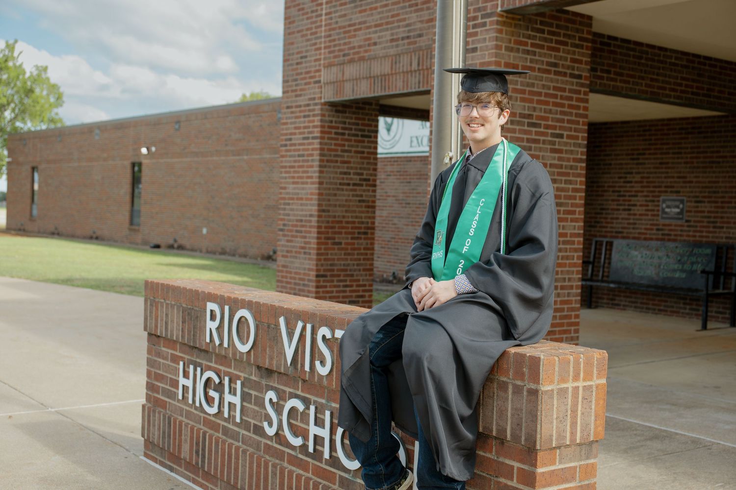 High school graduate in black cap and gown sitting on Rio Vista High School sign outside brick building.
