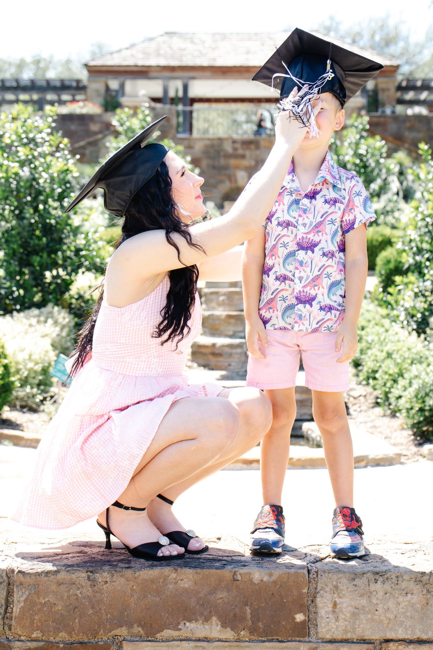 A mother in a pink dress adjusts a graduation cap on her young son outdoors in a garden.