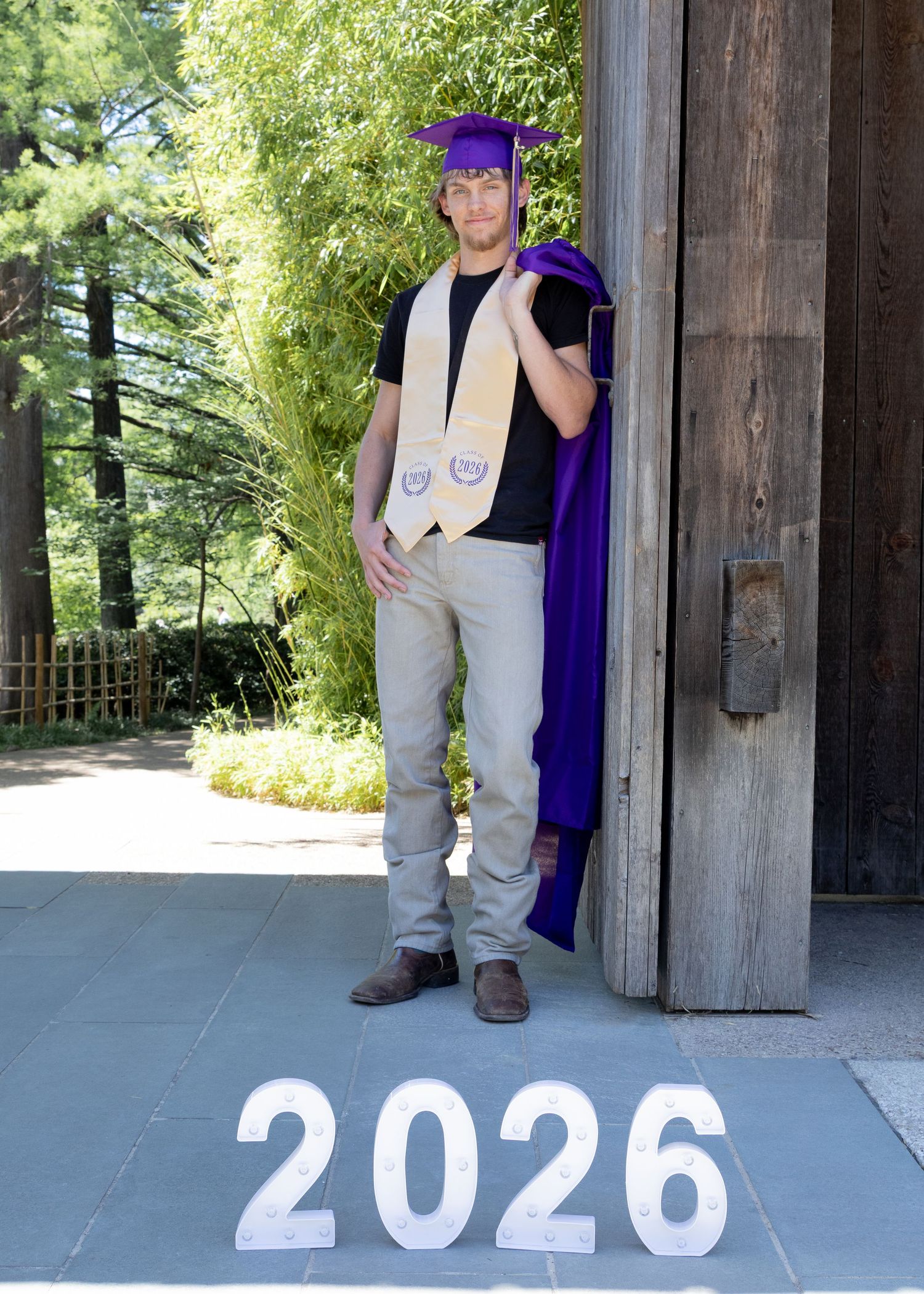 Young man in purple graduation cap and gown poses by wooden door with Class of 2026 numbers displayed.