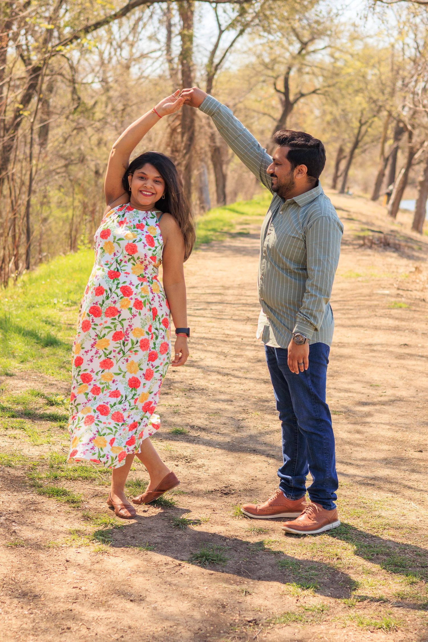 Joyful couple dancing on a scenic wooded path during a sunny outdoor maternity photoshoot.