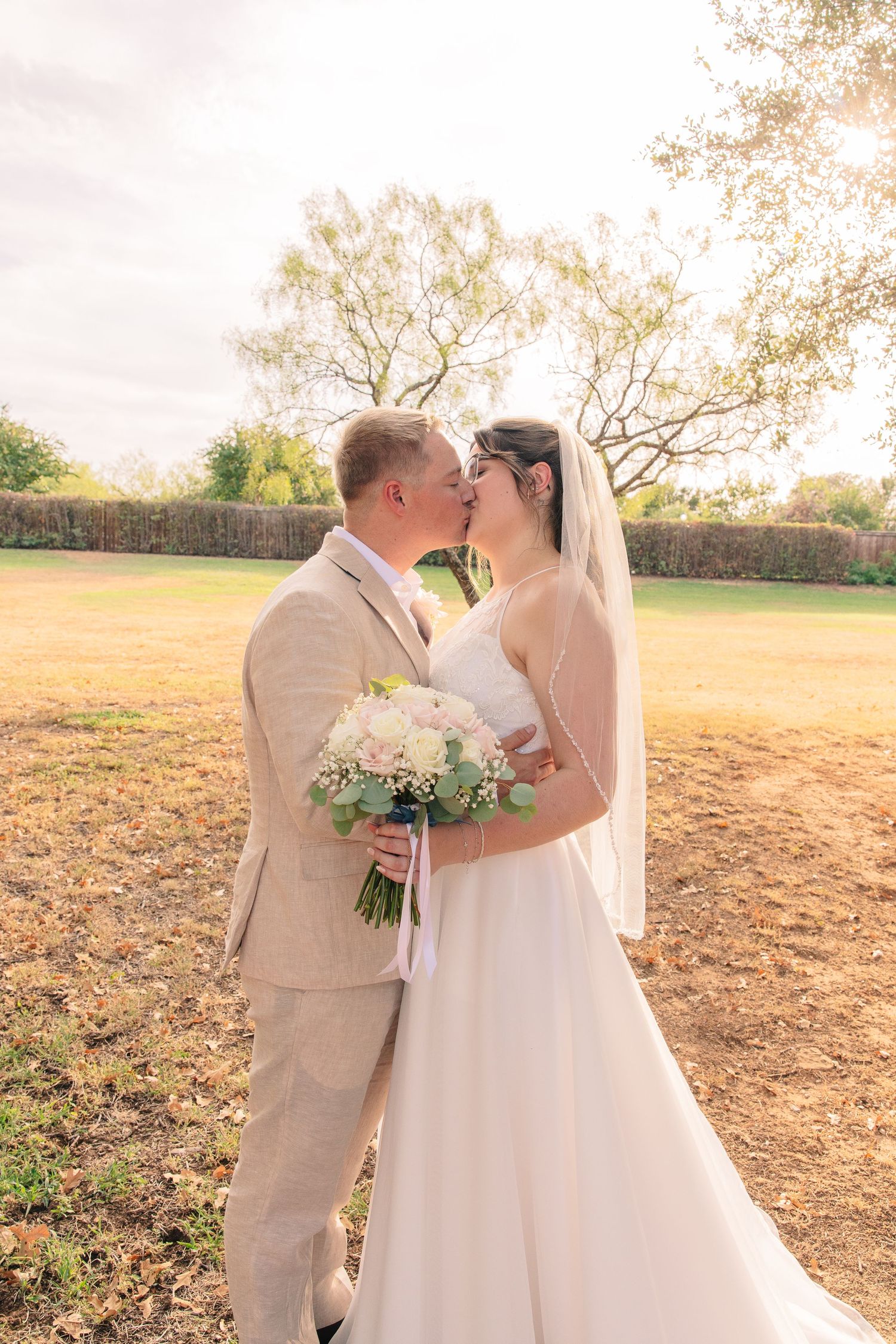 Bride in a white gown and groom in a tan suit share a kiss in a sunny field, holding a bouquet of white and green flowers.