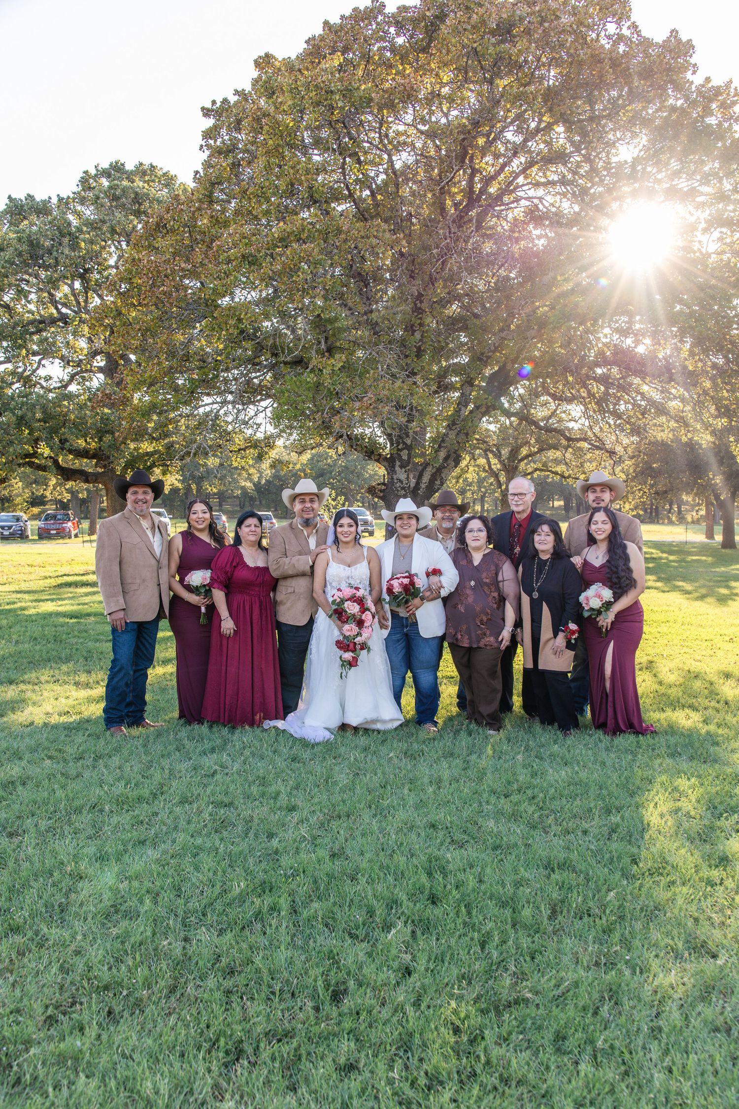 Bride in white gown and wedding party in burgundy dresses and cowboy hats pose on a grassy lawn under large oak trees at sunset.