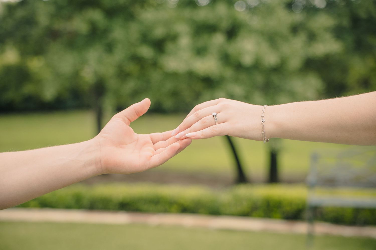 Two hands gently touching in a park, one wearing a diamond ring and bracelet, symbolizing love and connection.