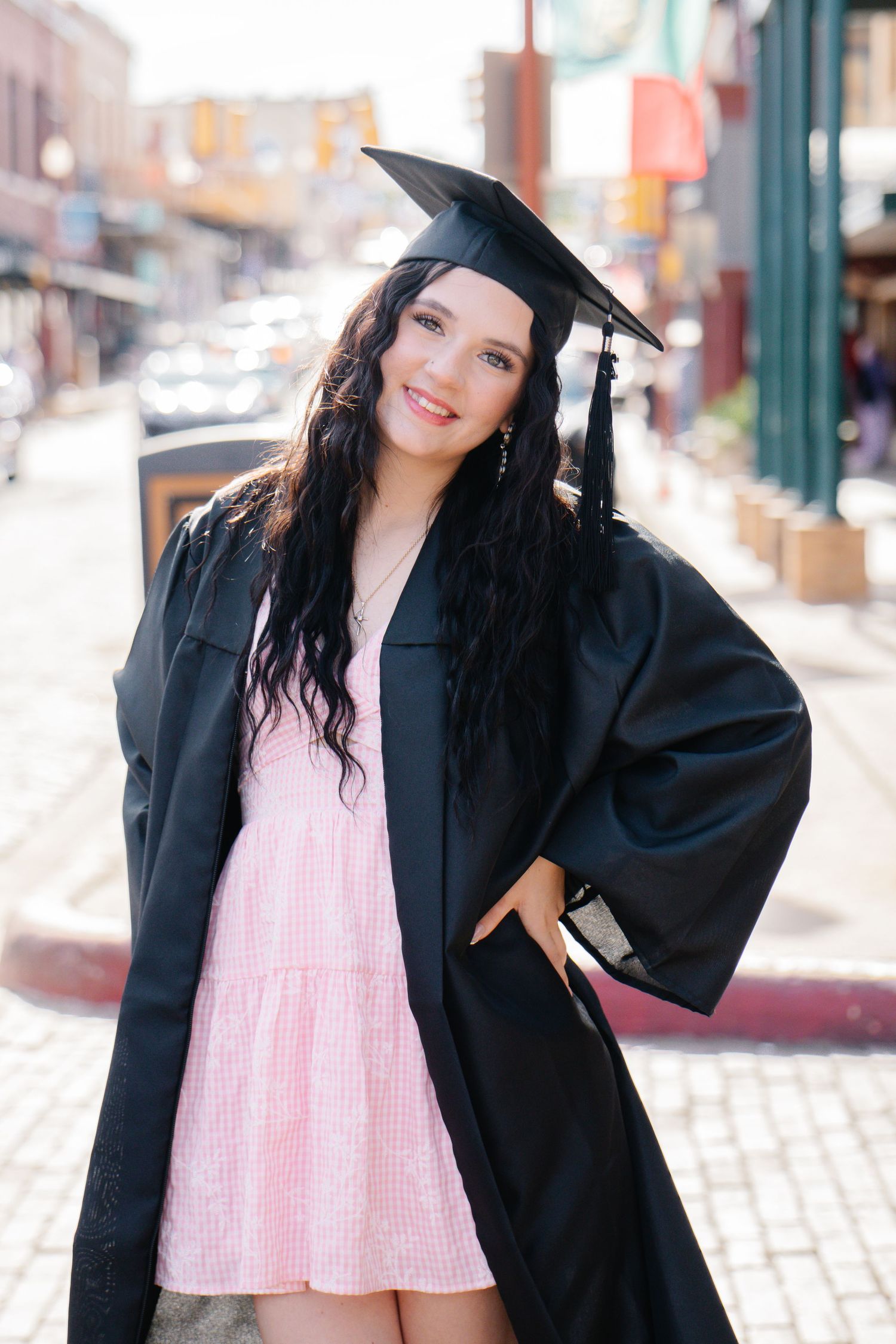 Smiling graduate in black cap and gown with pink dress posing confidently on cobblestone street.