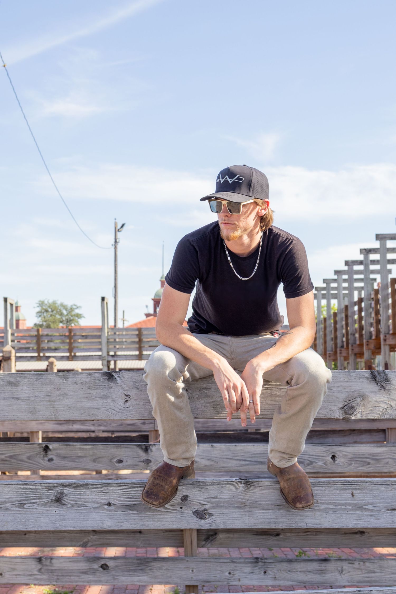 Young man in black shirt, khaki pants, and boots sitting on wooden fence outdoors.