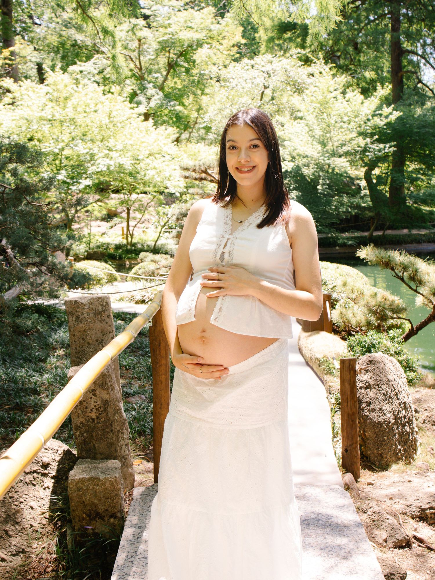 Pregnant woman in white dress holding baby bump while standing on bridge in Japanese garden.