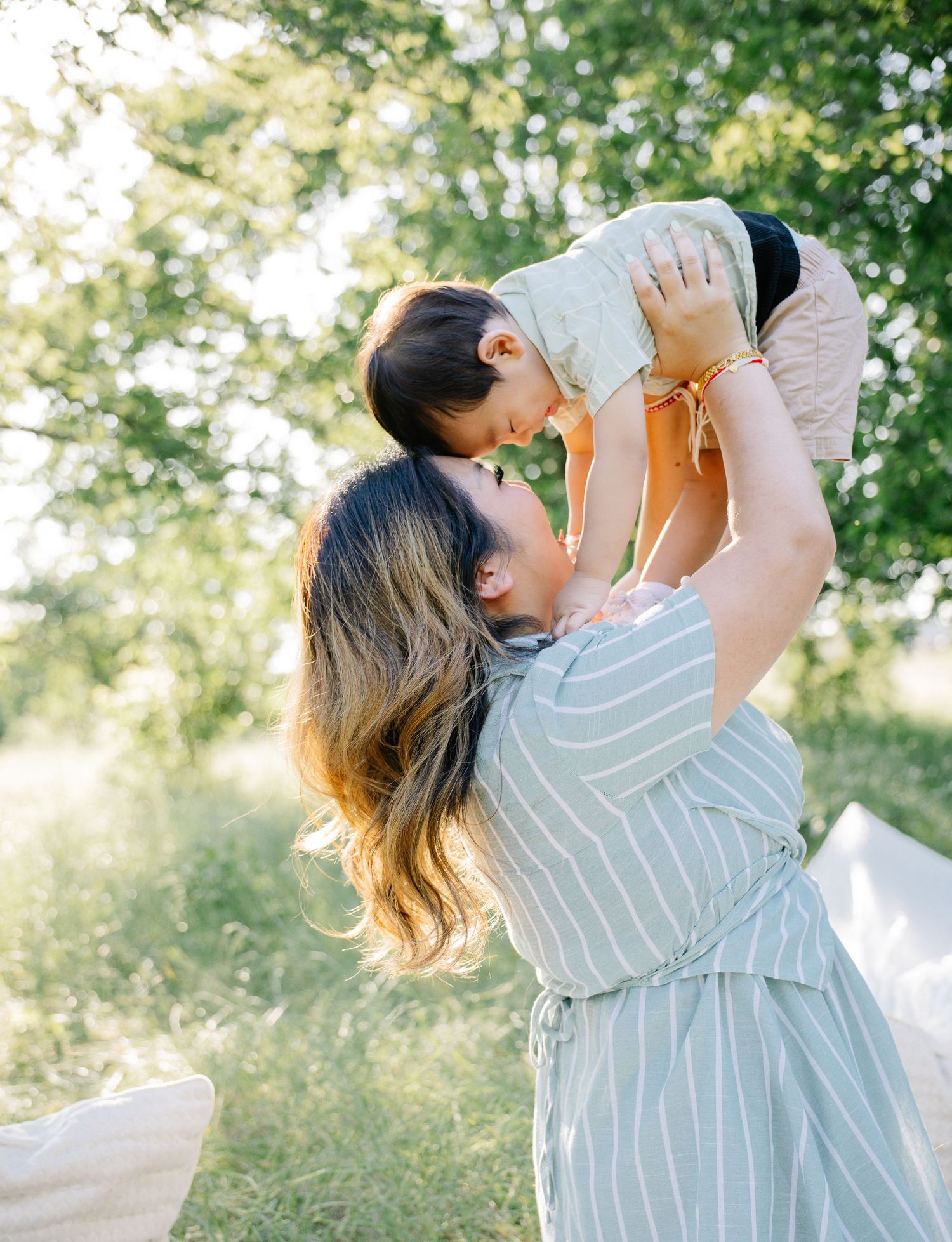 A mother joyfully lifts her toddler up in the air outdoors, surrounded by lush green trees in warm sunlight.