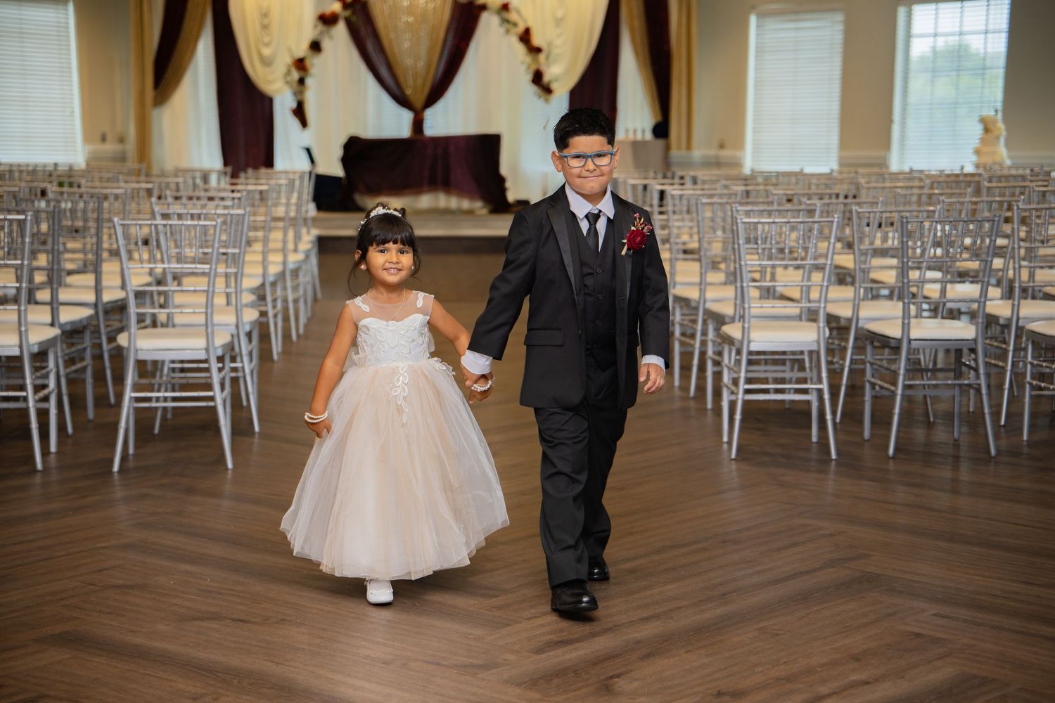 A young flower girl in a white dress walks hand-in-hand with a boy in a black suit down a wedding venue aisle.
