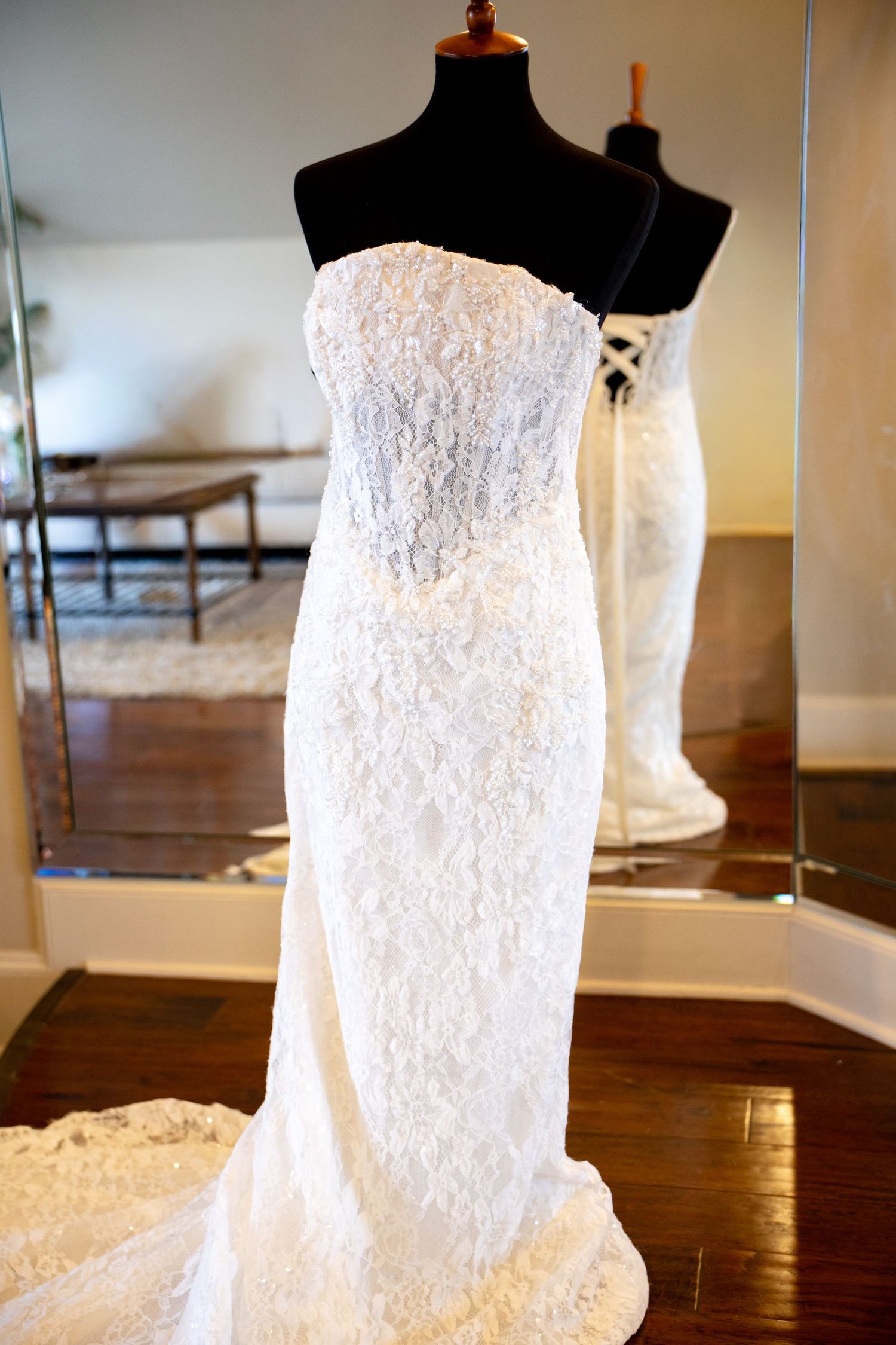 Elegant strapless white lace wedding gown displayed on a black mannequin in a bridal boutique with mirror reflection.
