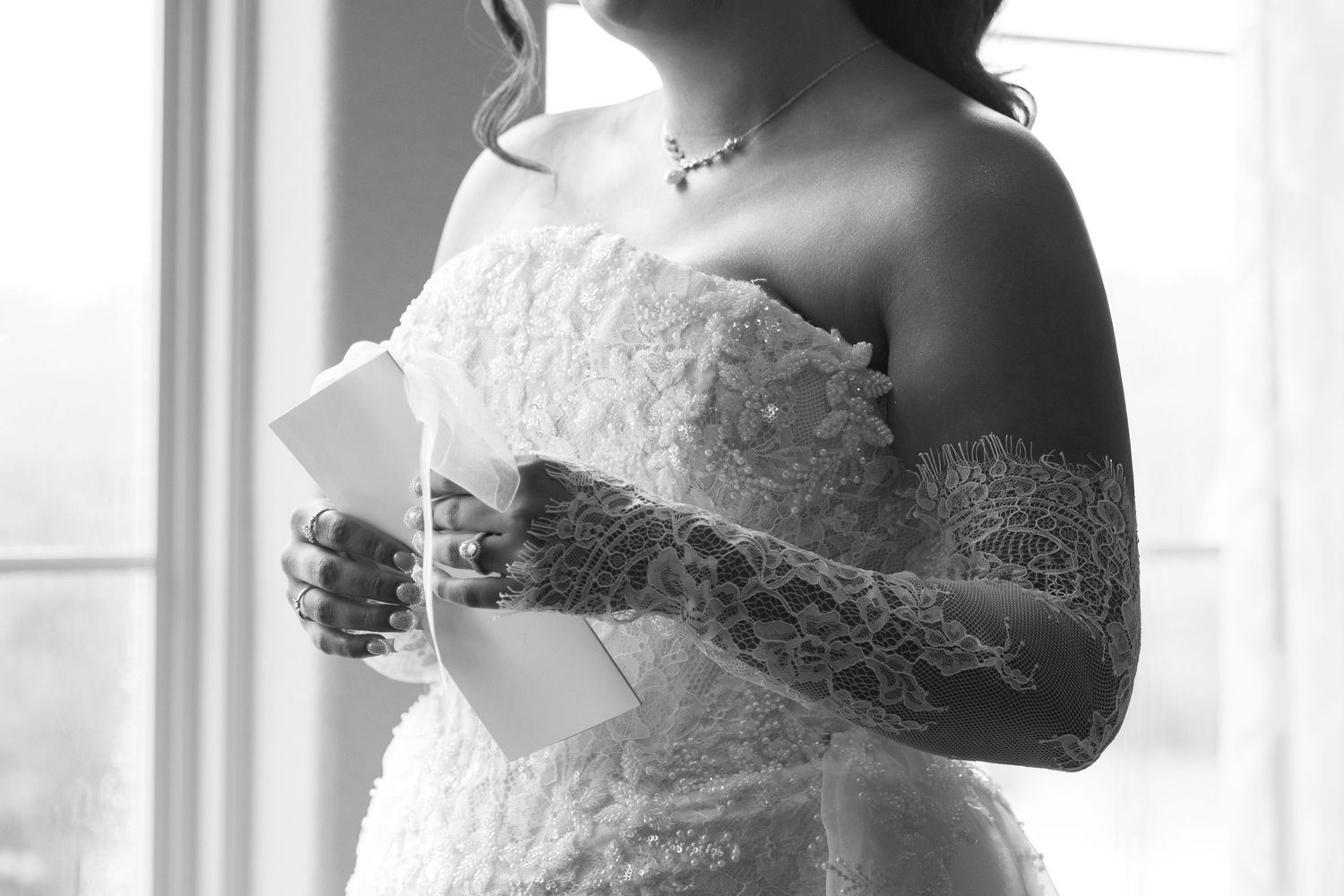 Bride in off-shoulder lace wedding gown reading a letter by window, black and white bridal portrait.