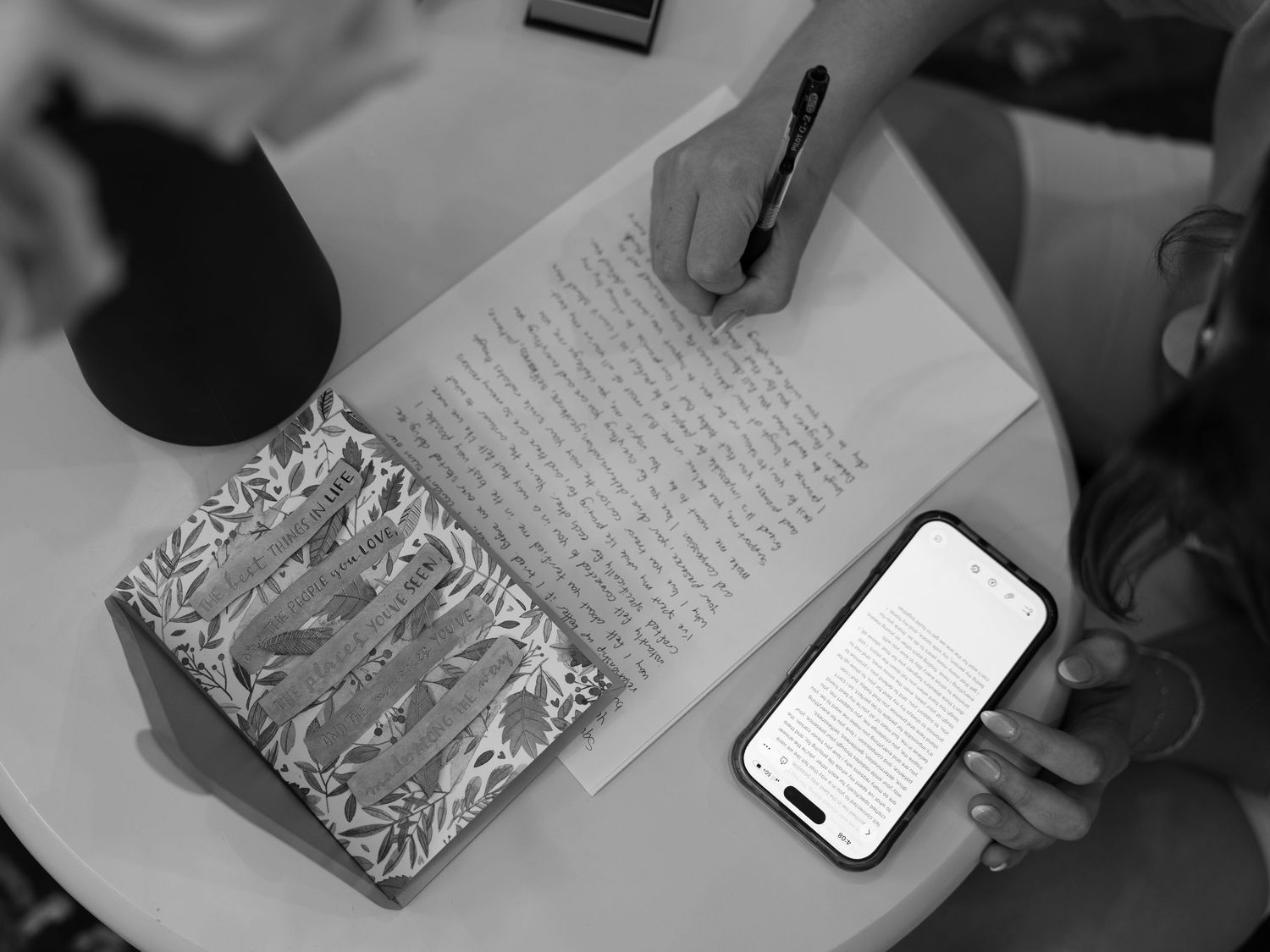 Overhead black and white view of a person handwriting a letter on lined paper beside a patterned notebook and a smartphone on a round table.