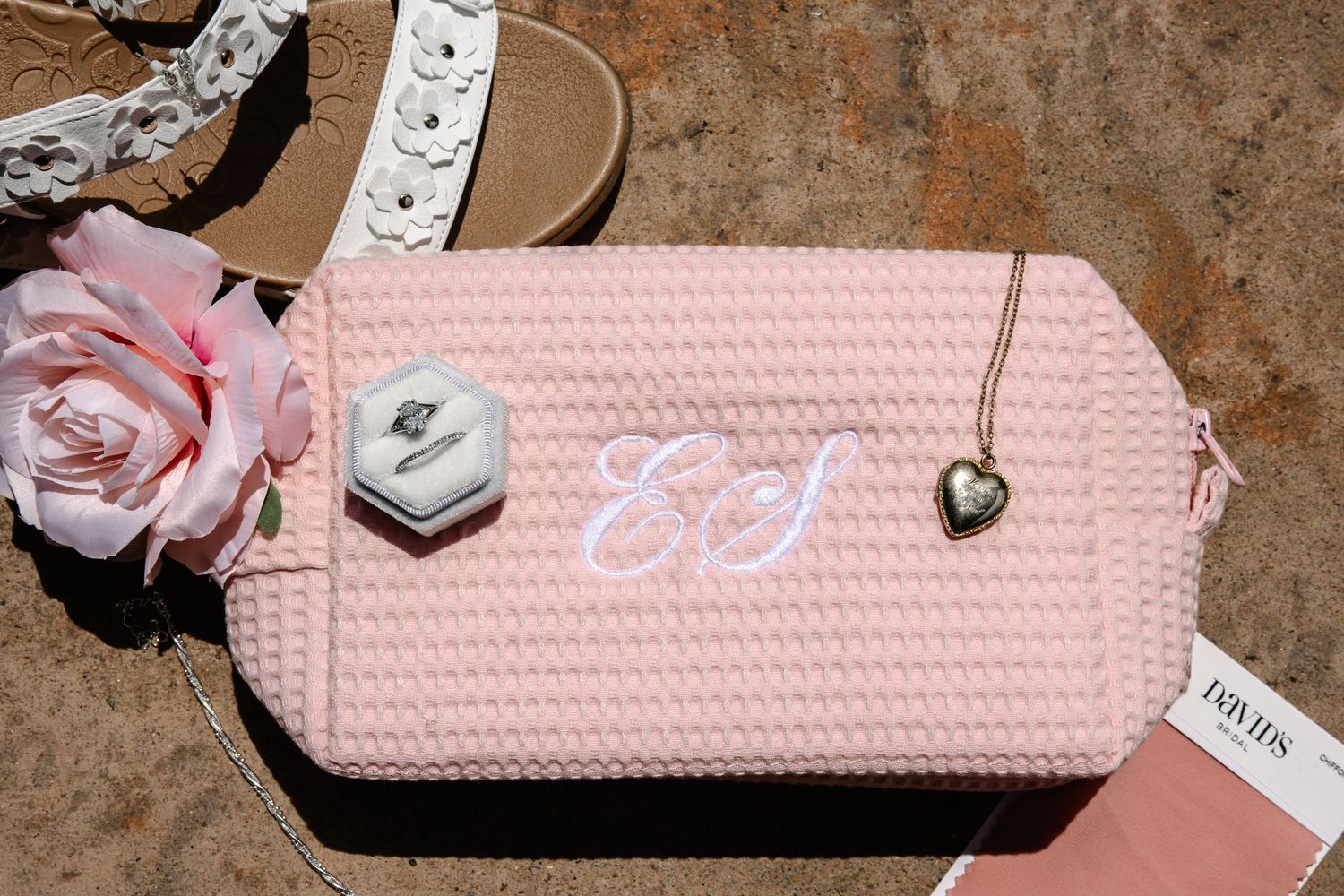 Pink waffle-textured makeup bag with white monogram, a heart locket, pink rose, ring box and sandals on a sunlit stone surface.