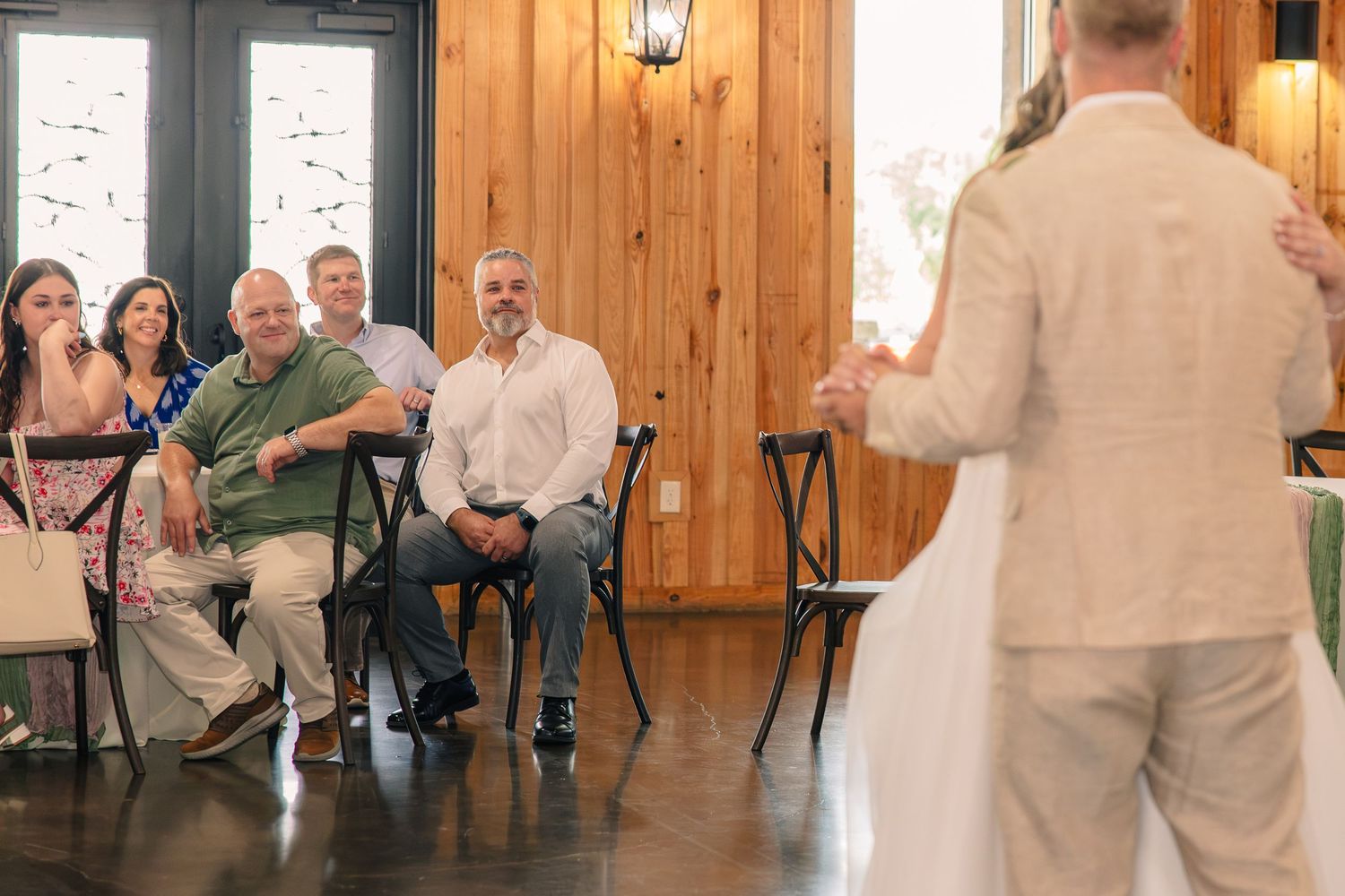 Wedding guests smile from their seats inside a rustic wood-paneled venue while the bride and groom stand in the foreground.