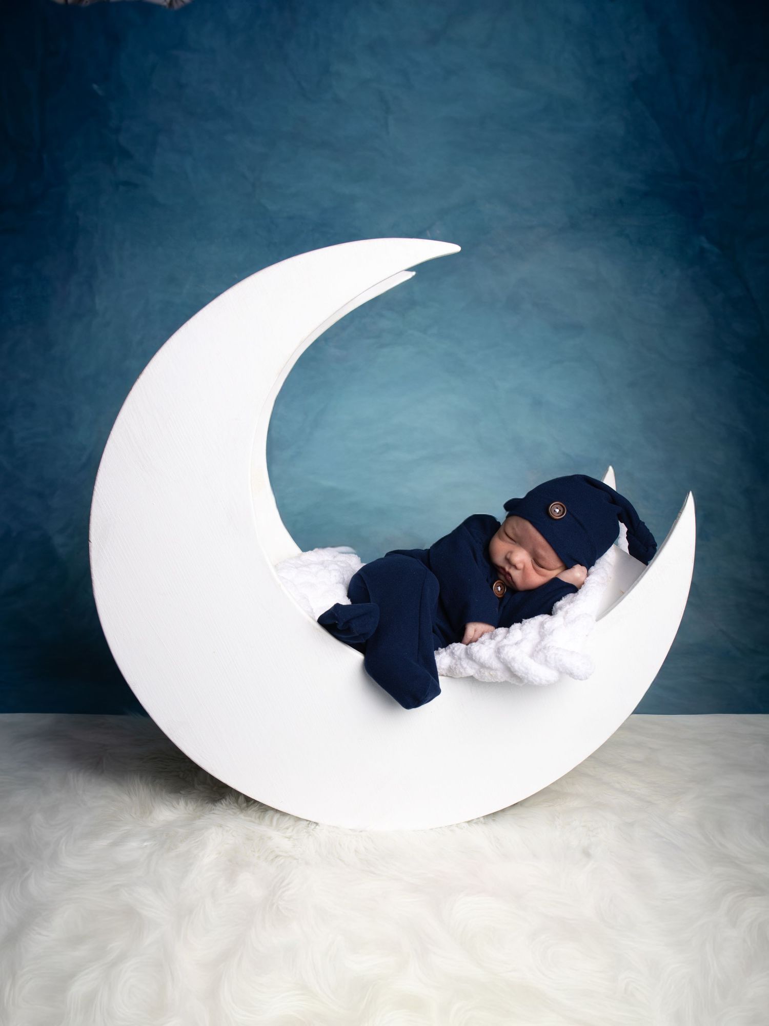 Newborn baby in a navy outfit and beanie sleeping on a white crescent moon prop with a soft blanket against a blue backdrop. Haslet Texas