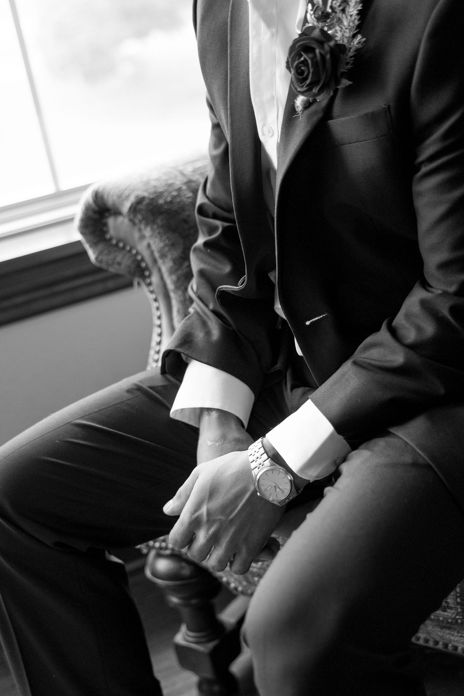 Groom in black suit with boutonniere and watch sits in chair, black and white wedding photo.