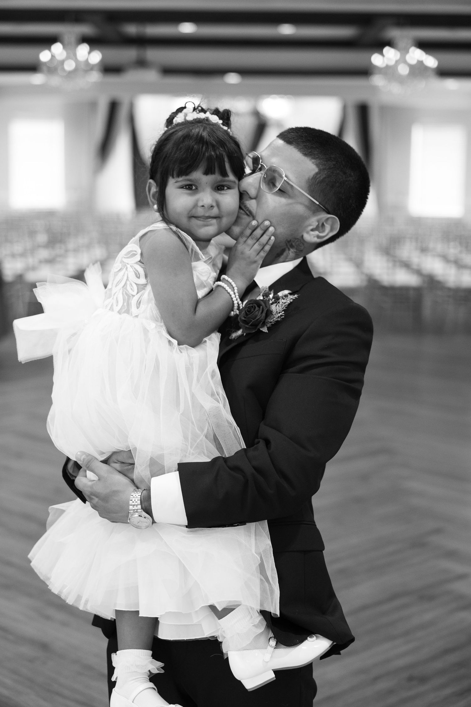 Groom in black tuxedo tenderly kisses young flower girl in white dress at elegant wedding venue.
