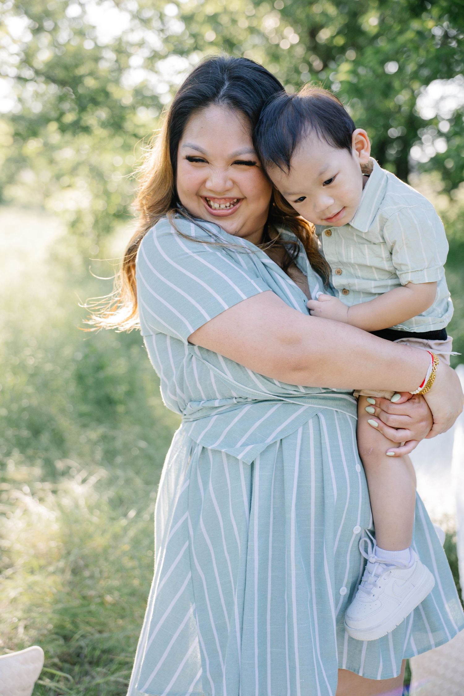 A smiling woman in a striped dress holds a young toddler boy outdoors in a sunlit green park setting.