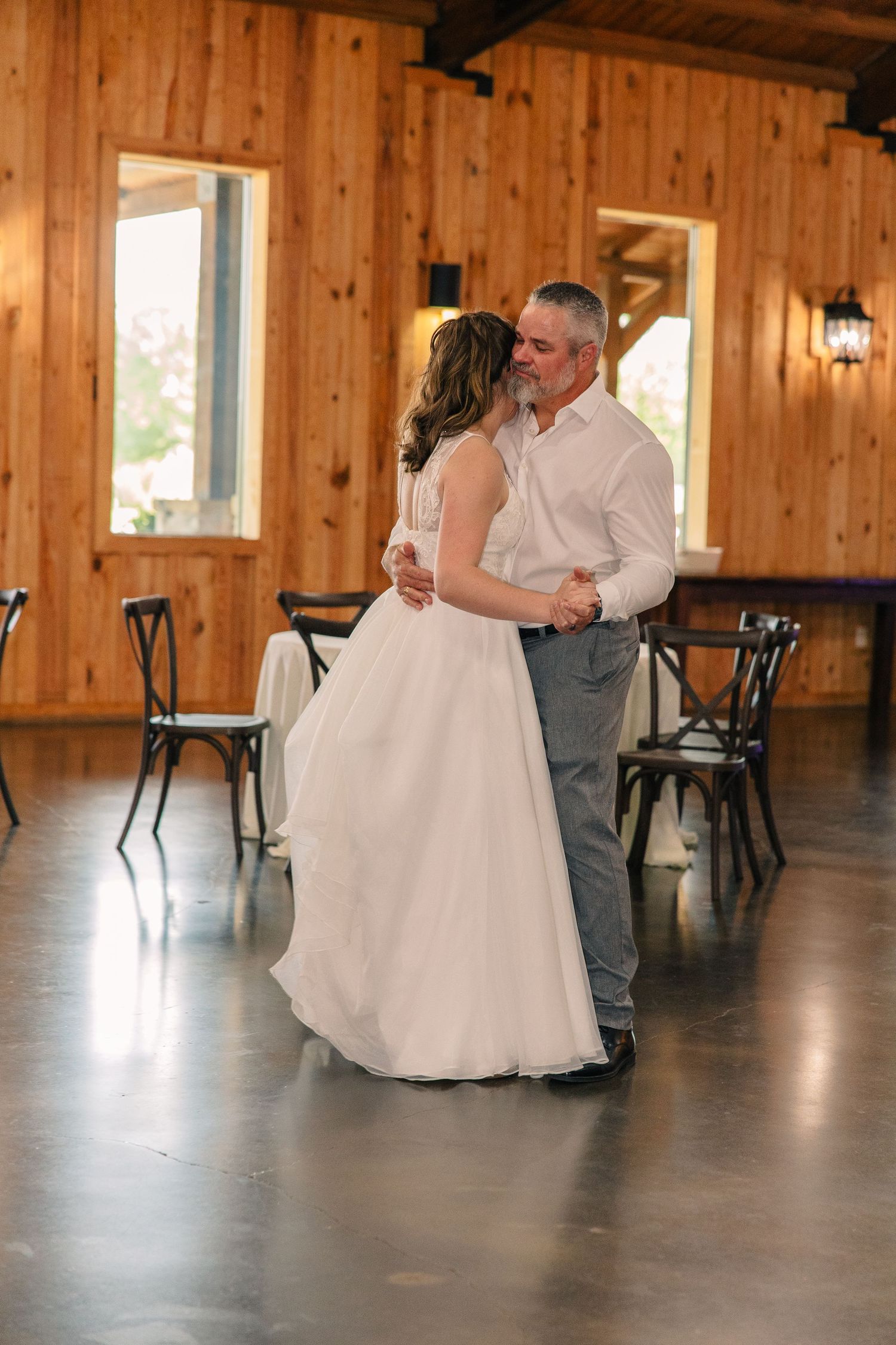 Bride dances with her father in a white gown and gray pants on a polished floor in a rustic barn wedding venue.