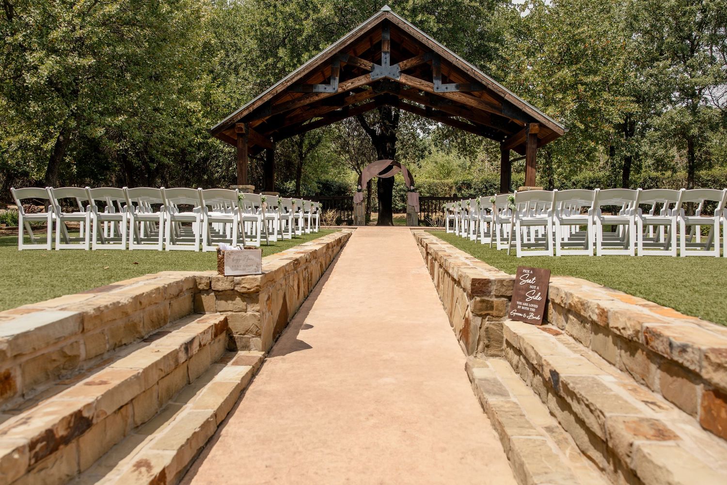 Rustic wooden outdoor wedding pavilion with a stone aisle and white folding chairs set among tall trees.