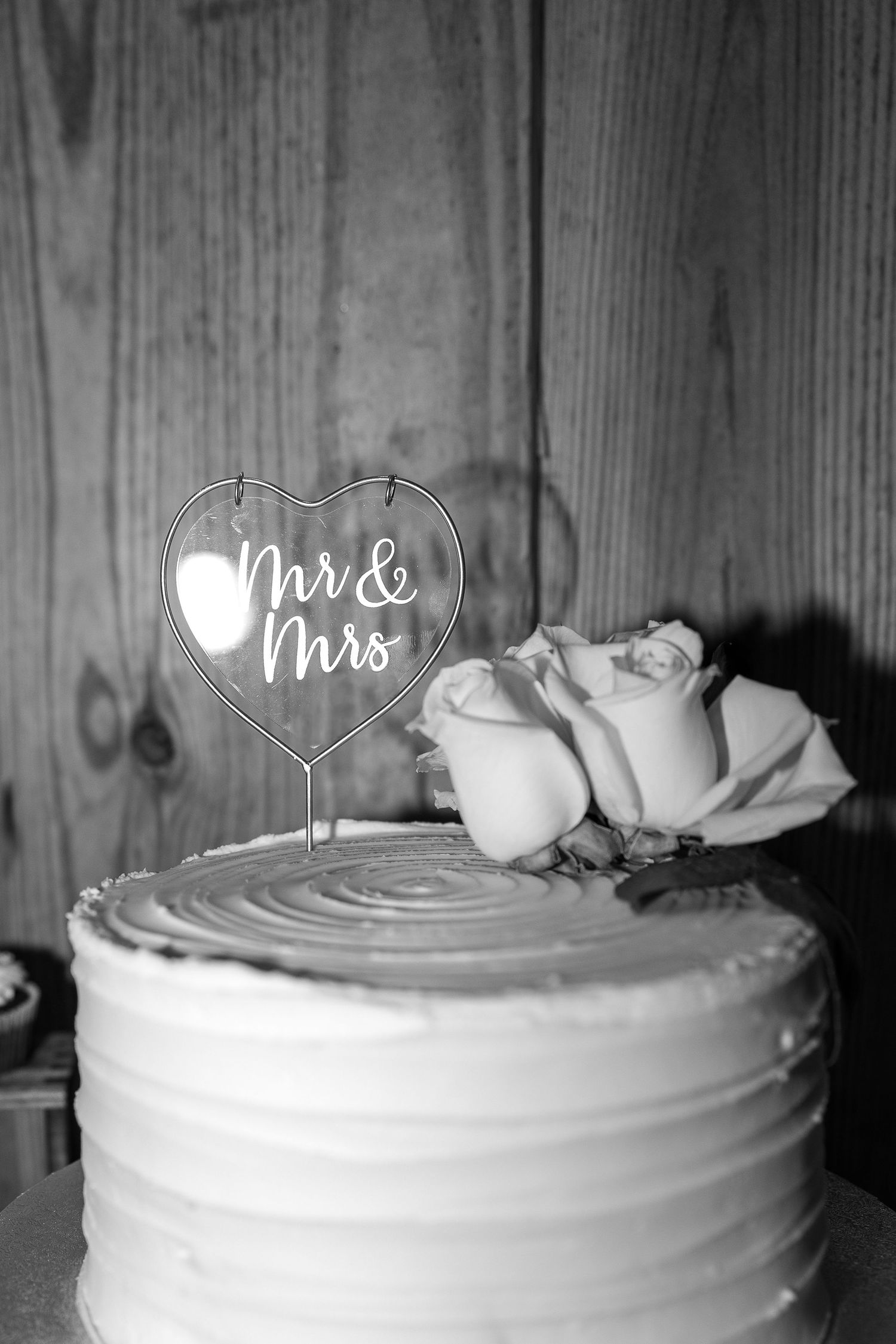 Black and white photo of a buttercream wedding cake topped with a heart-shaped Mr & Mrs cake topper and fresh roses.