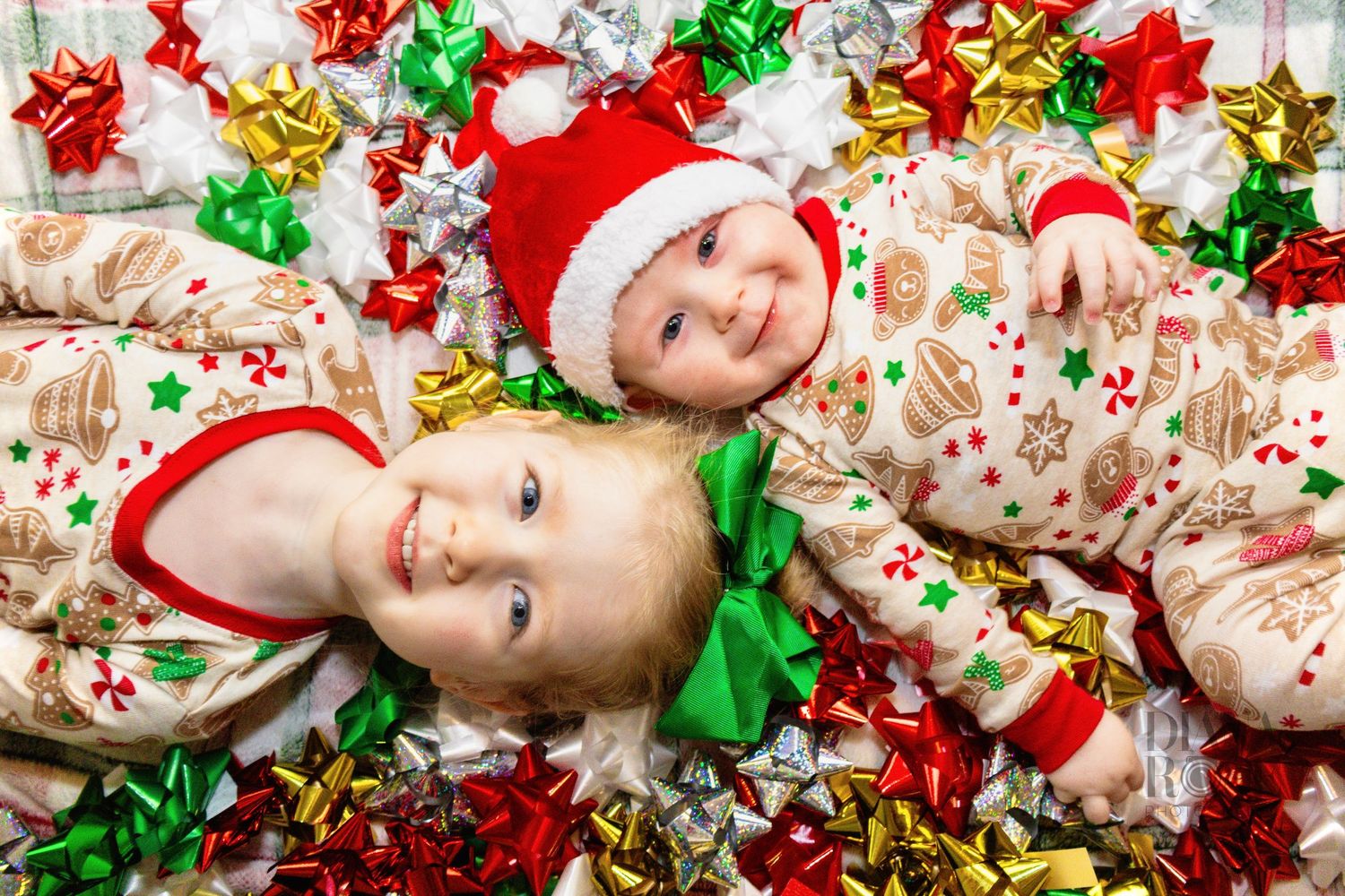 Two smiling children in matching Christmas pajamas, one wearing a Santa hat, lie surrounded by red, green, and gold holiday bows. Haslet Tx