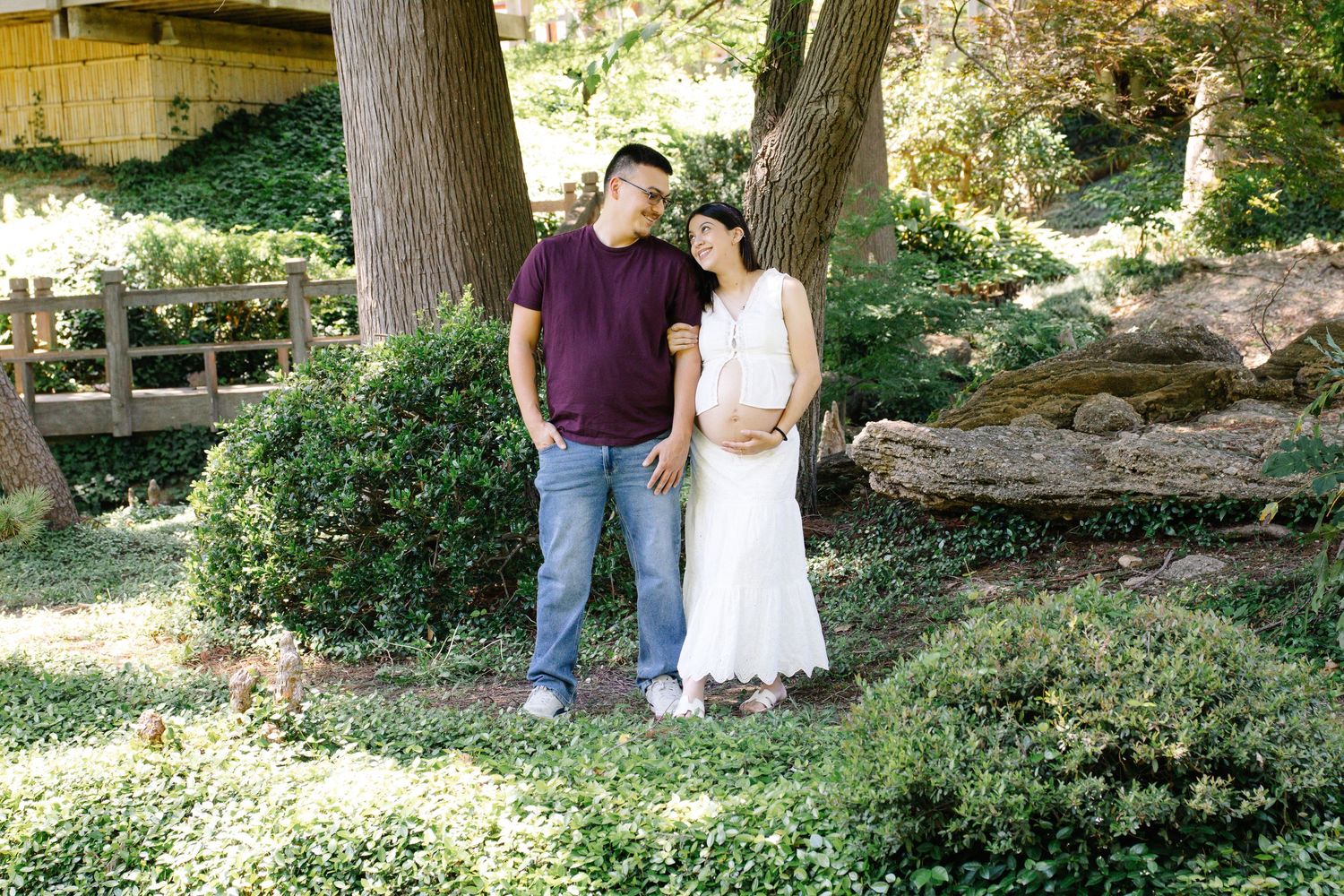 Expecting couple standing together in a lush garden, woman in white dress cradling baby bump.