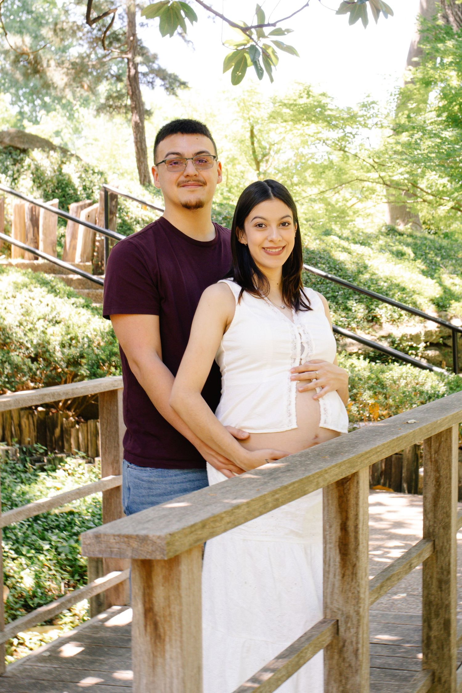 Happy expecting couple embracing on wooden bridge surrounded by lush green trees in park.