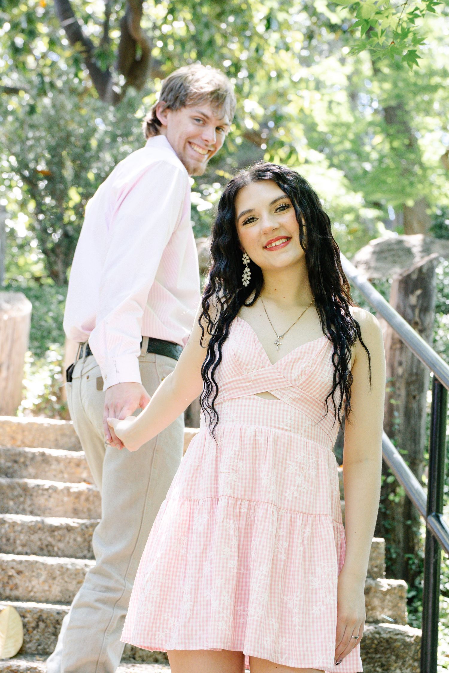 Smiling couple holding hands on stone steps outdoors, woman in pink dress and man in white shirt.