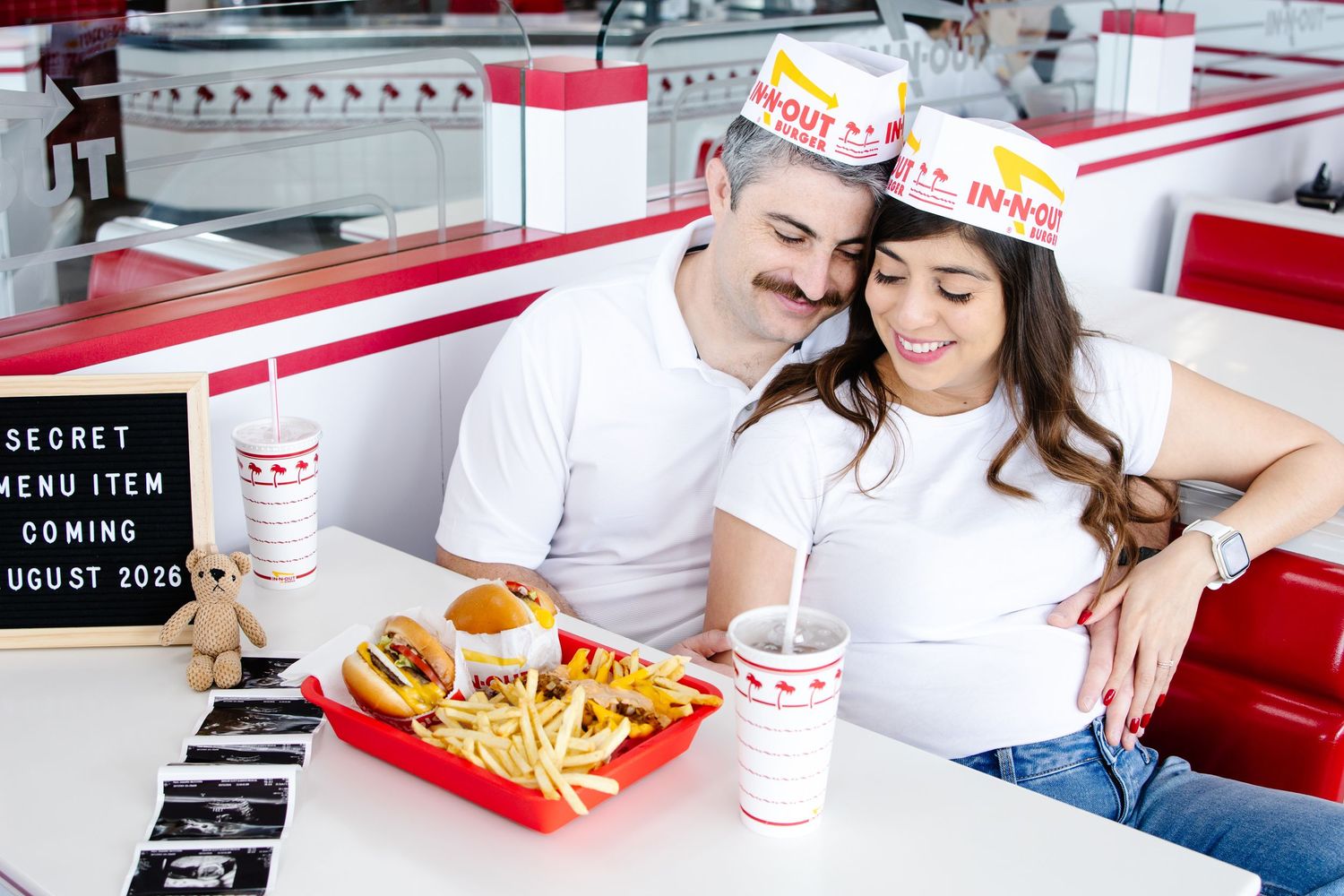 Pregnant couple wearing In-N-Out hats sitting at diner table with burgers and pregnancy announcement sign.