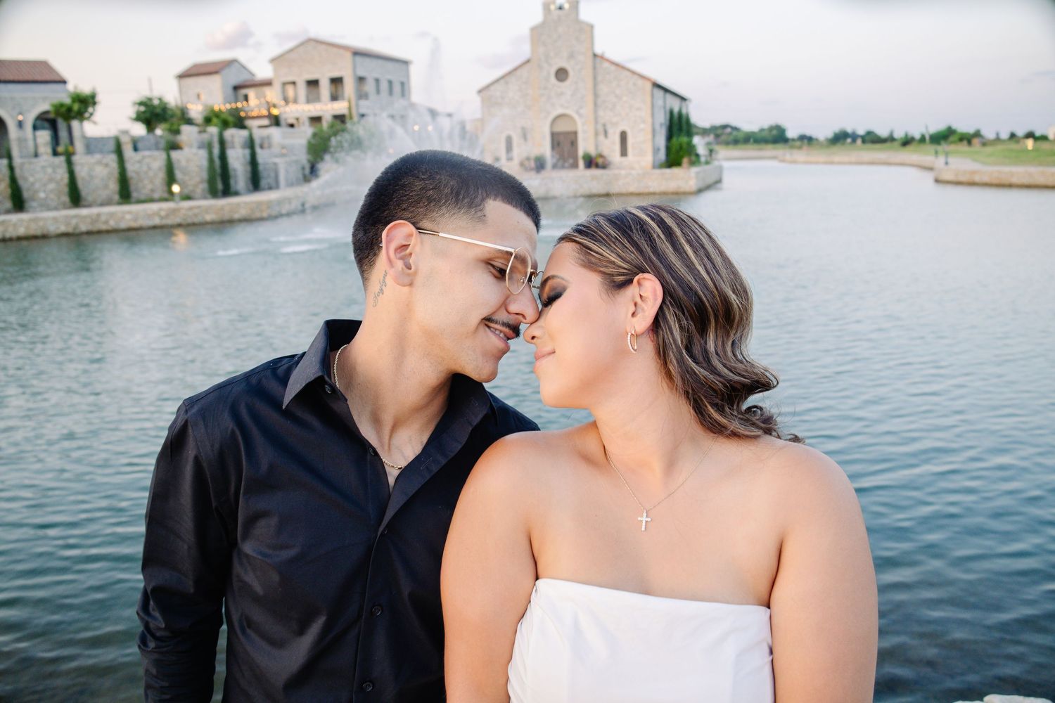 Couple in romantic pose near a lakeside chapel, man in black shirt and woman in white strapless dress touching foreheads.