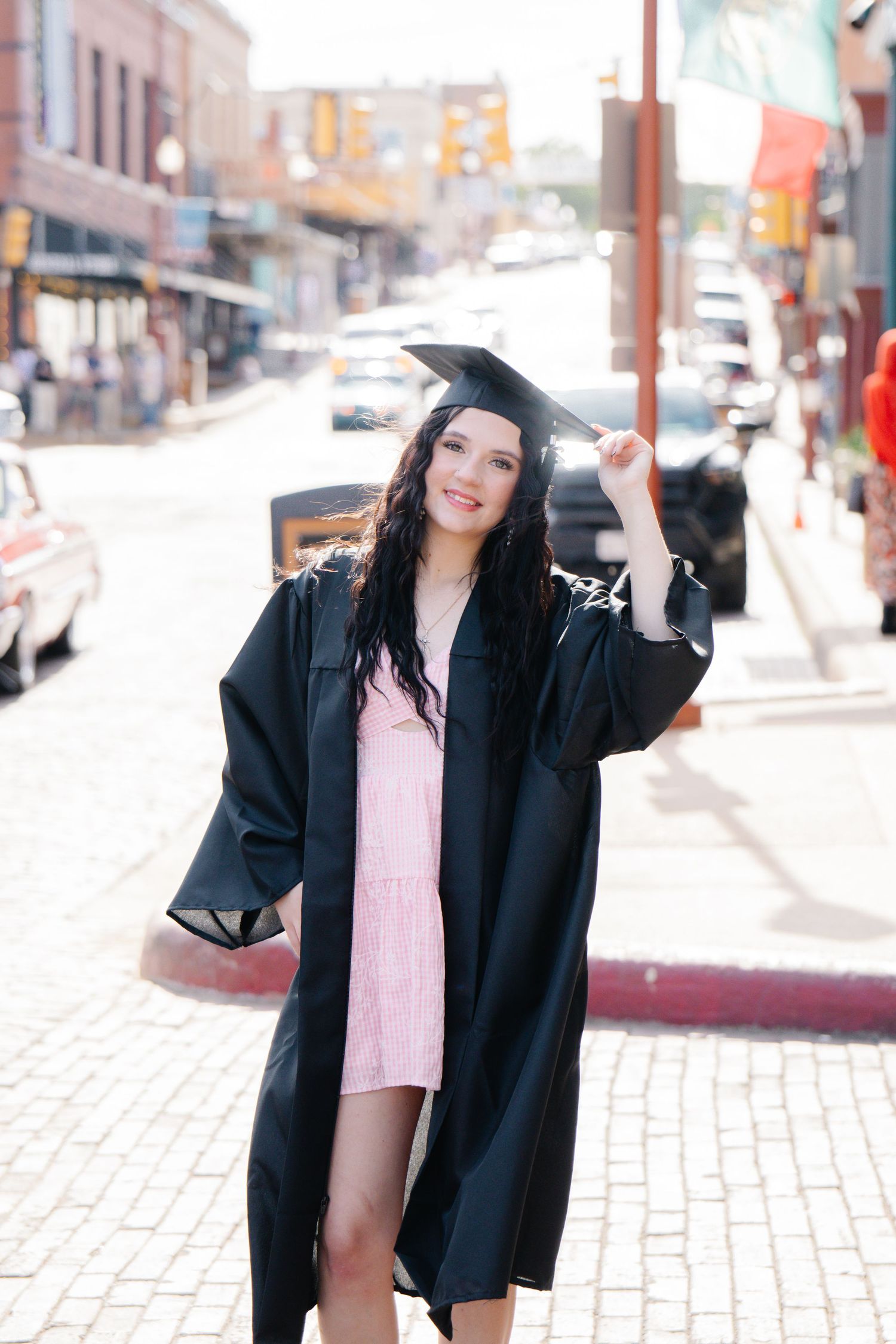 Young woman in graduation cap and gown standing on sunny brick street, holding cap tassel.