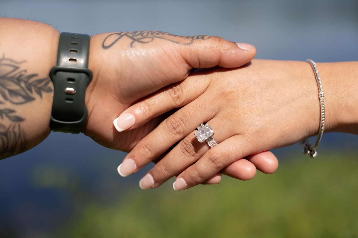 Close-up of couple holding hands showing diamond engagement ring, one hand with tattoos and smartwatch.