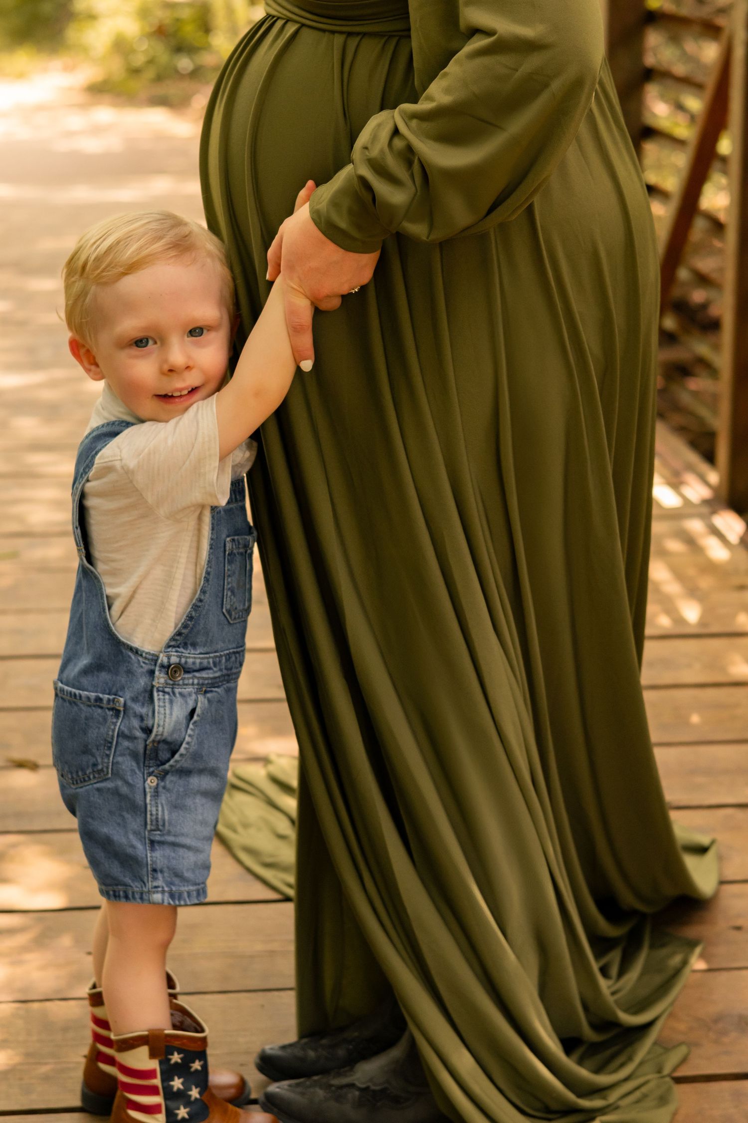 Toddler boy in denim overalls holding hands with his pregnant mother in an olive green maternity gown.