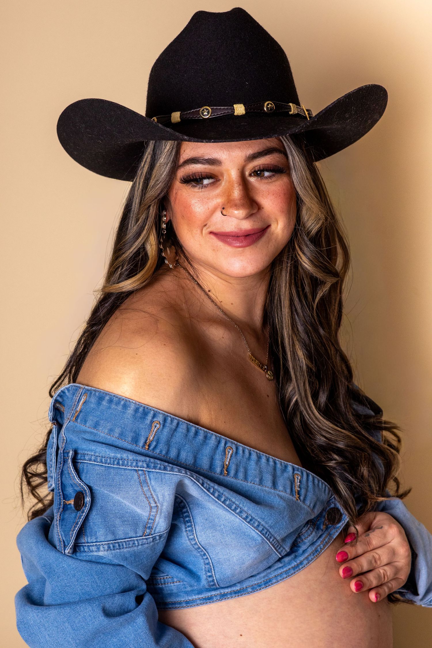 Smiling woman in black cowboy hat and off-shoulder denim top posing against a warm beige background.