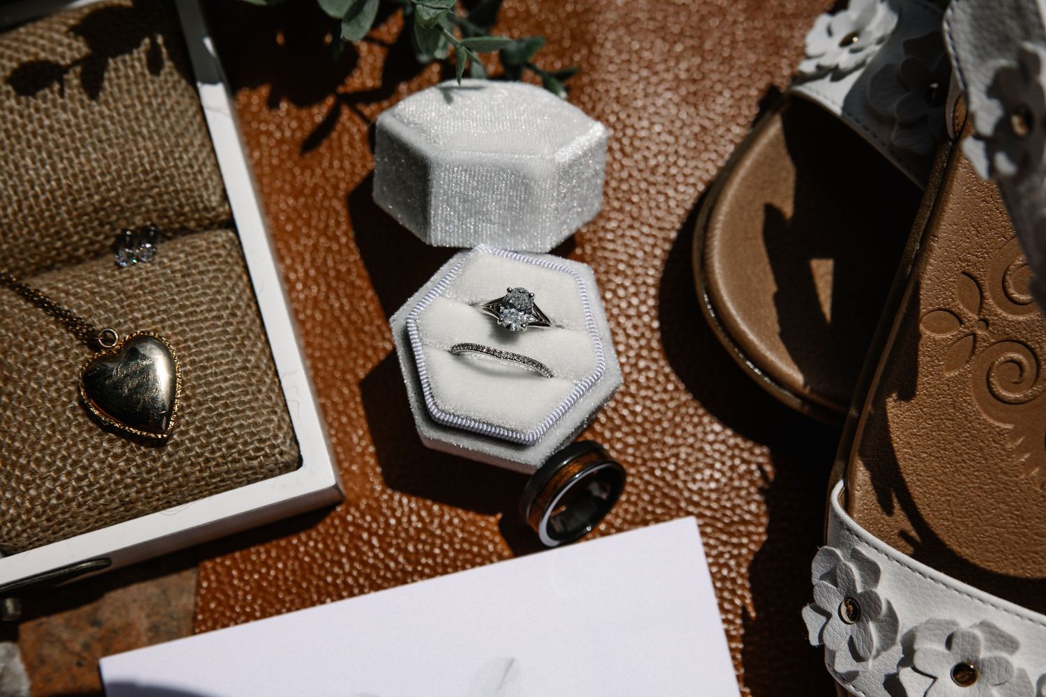 Wedding flat lay with a diamond ring in a white velvet box, a heart locket necklace, a ring box, and floral sandals on leather.
