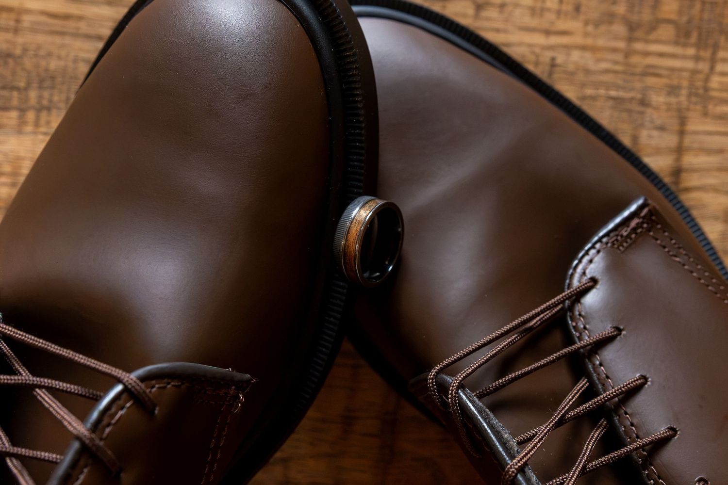 Close-up of a pair of dark brown leather lace-up shoes with a round metal ring accent, resting on a rustic wooden surface.