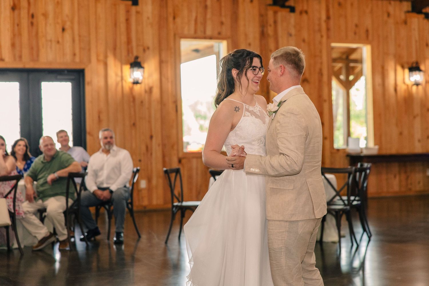 Bride and groom share a first dance in a rustic wooden barn venue while seated wedding guests watch from nearby tables.