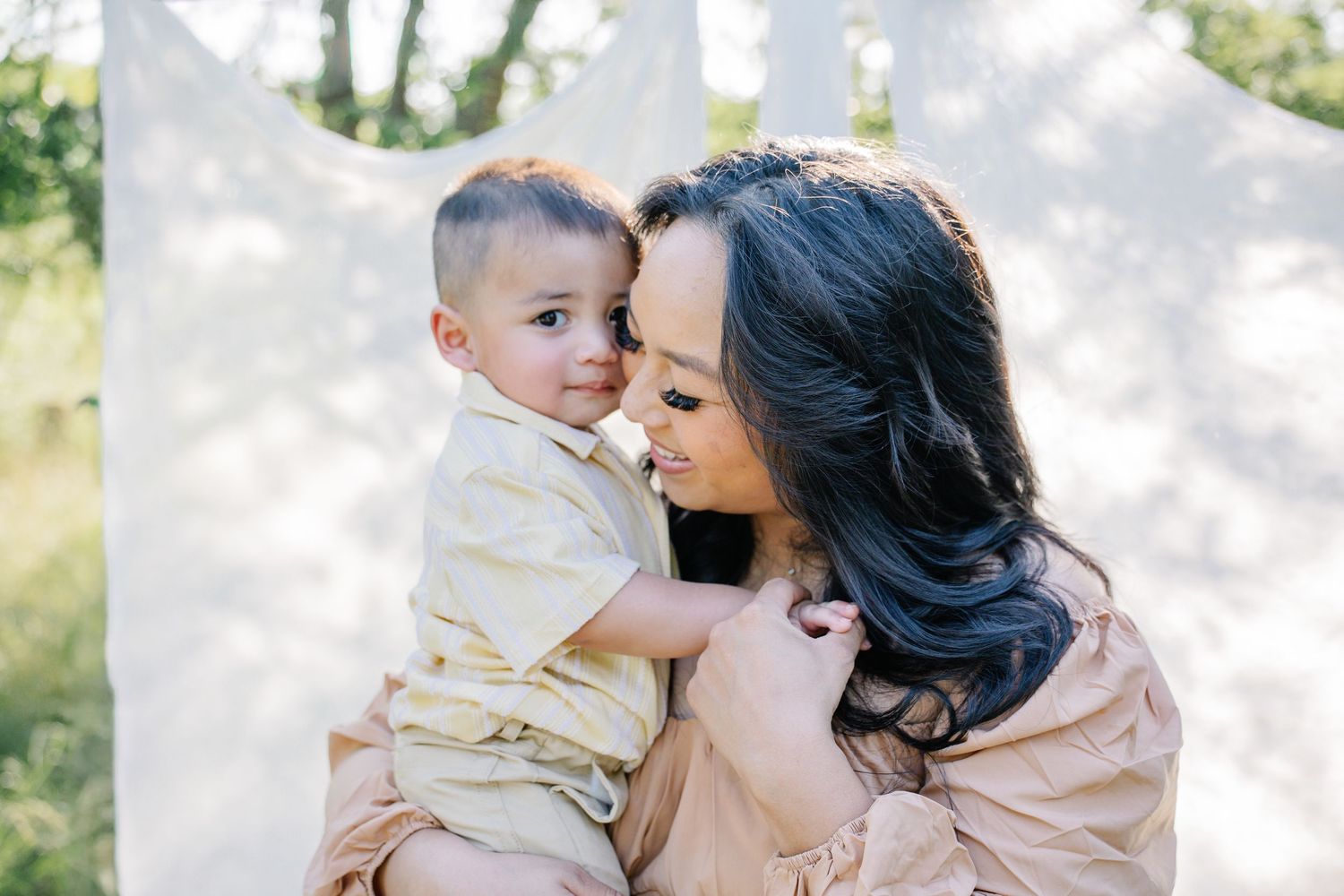 A mother tenderly holds her baby boy outdoors, sharing a sweet moment against a soft white backdrop.