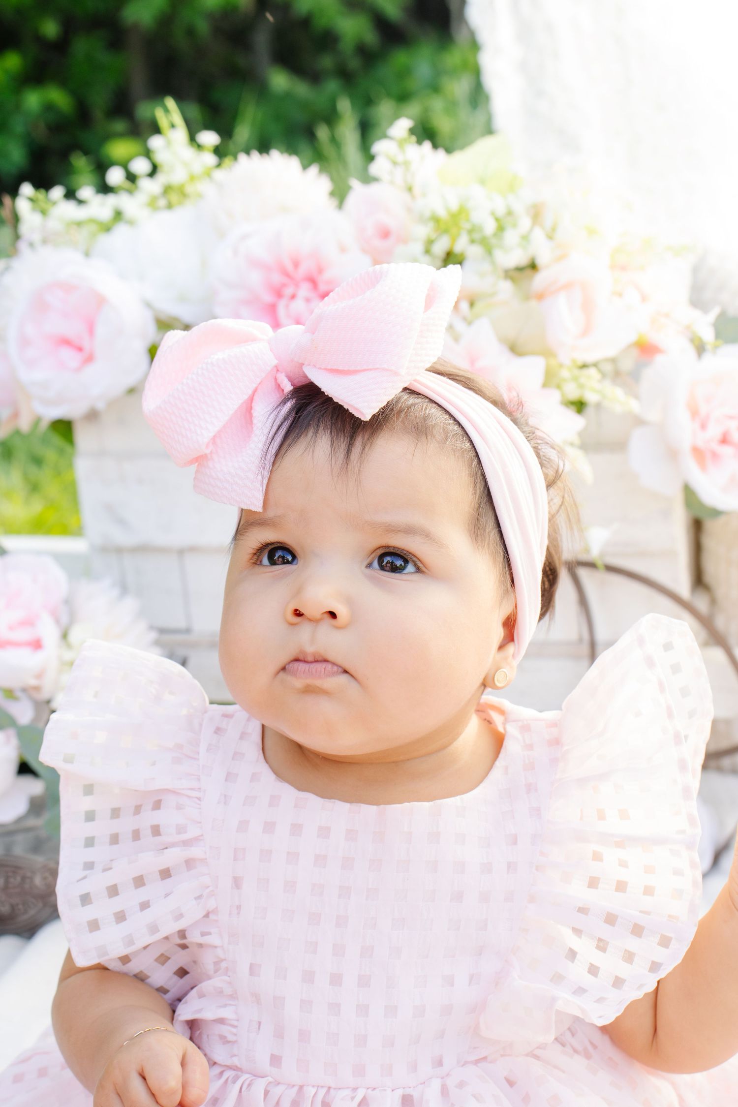 Adorable baby girl in pink dress and bow headband sits outdoors surrounded by soft pink flowers.