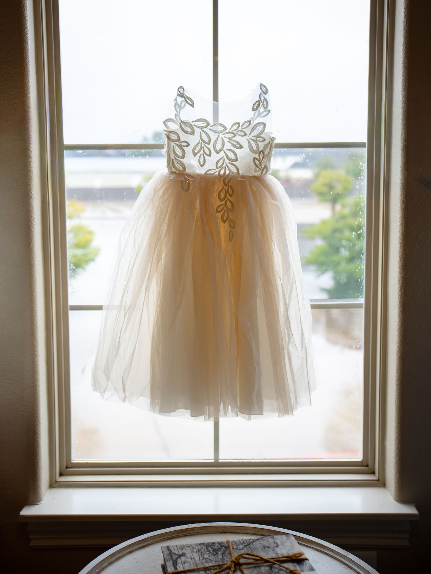 Delicate flower girl dress with gold embroidery and tulle skirt hanging in a sunlit window.