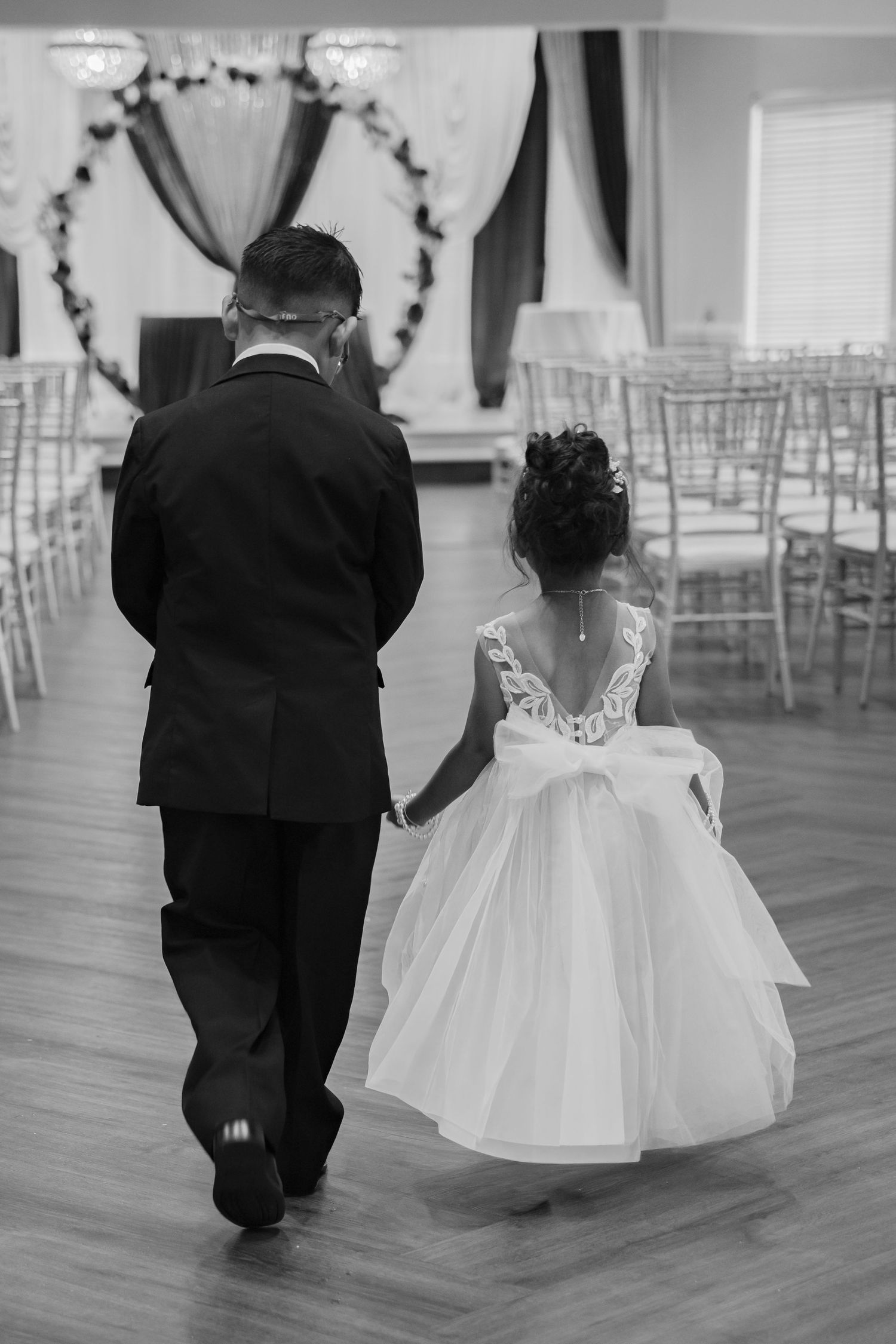 Black and white photo of a man in a suit walking hand in hand with a flower girl in a white dress at a wedding reception.