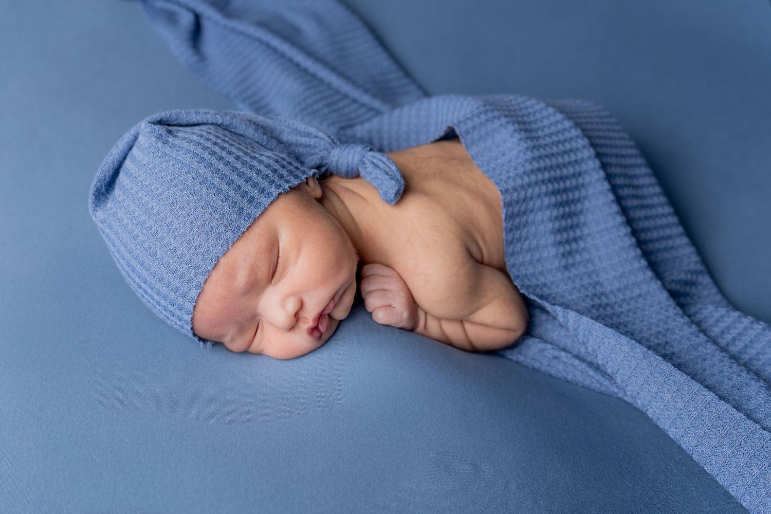 Newborn baby sleeping peacefully wrapped in a soft blue knit blanket and matching hat on a blue background.