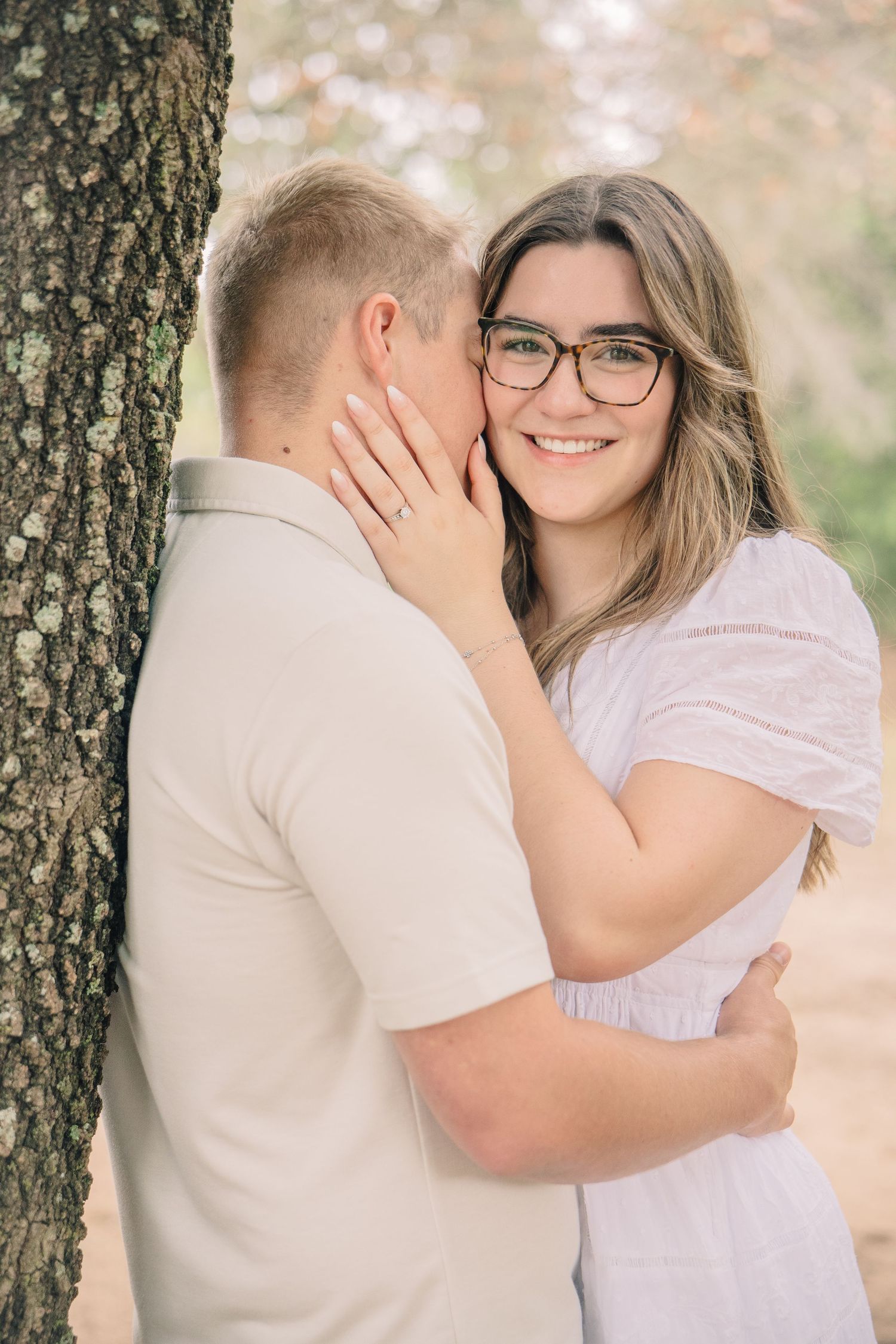 A smiling woman with glasses in a white dress embraces a man near a tree in an outdoor engagement photo.