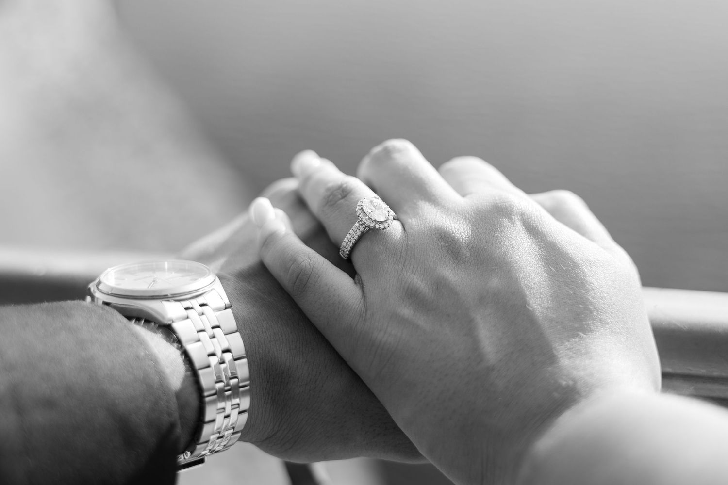 Black and white photo of couple holding hands, woman wearing diamond engagement ring, man wearing silver watch.